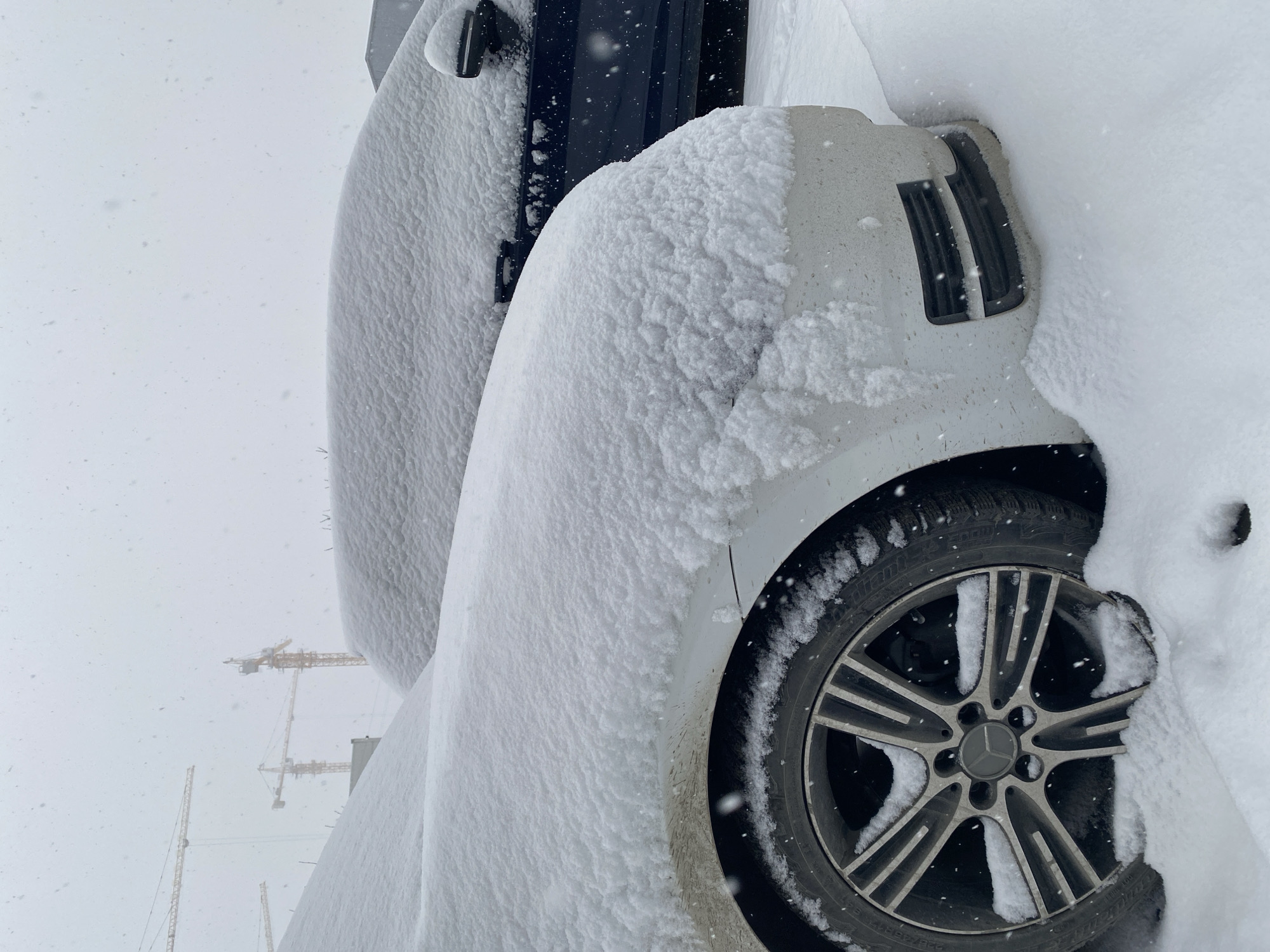 The image shows a white car heavily covered in snow, with only a portion of the front wheel and tire visible. The car is parked outdoors in what appears to be a snowy environment. In the background, indistinct shapes of construction cranes are visible. No people are visible in the image. The location is identified as Yekaterinburg, Russia.