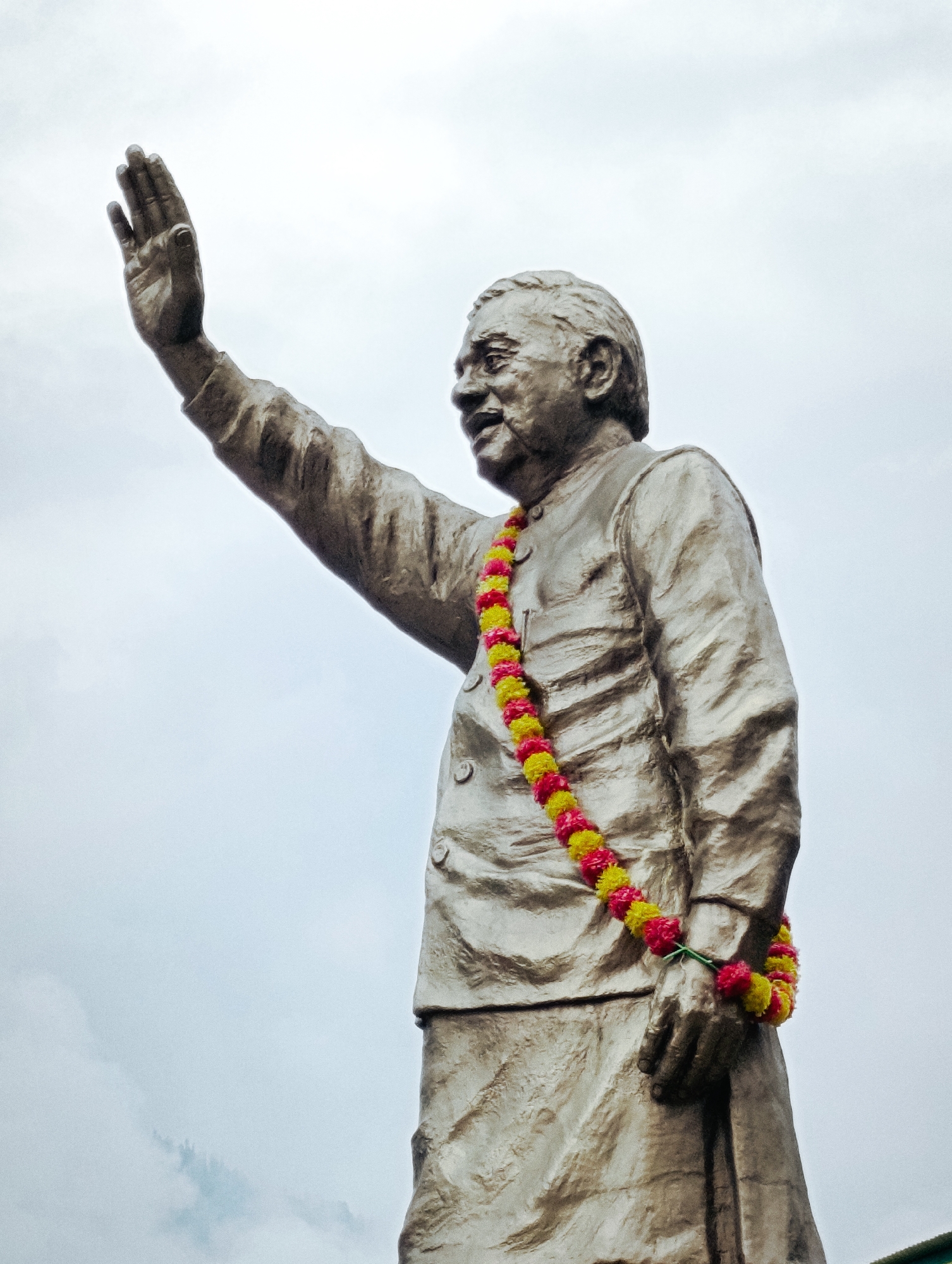 A low-angle, medium shot captures a bronze statue of a man against an overcast sky. The statue depicts the man with his right arm raised and palm facing outward, as if waving or giving a blessing. A garland of yellow and pink flowers adorns his neck and drapes over his raised arm and chest. The man is wearing traditional Indian attire, including a jacket with buttons and a dhoti or similar lower garment. The statue is situated in the city of Manali, India, a popular tourist destination in the Himalayas known for its scenic beauty.