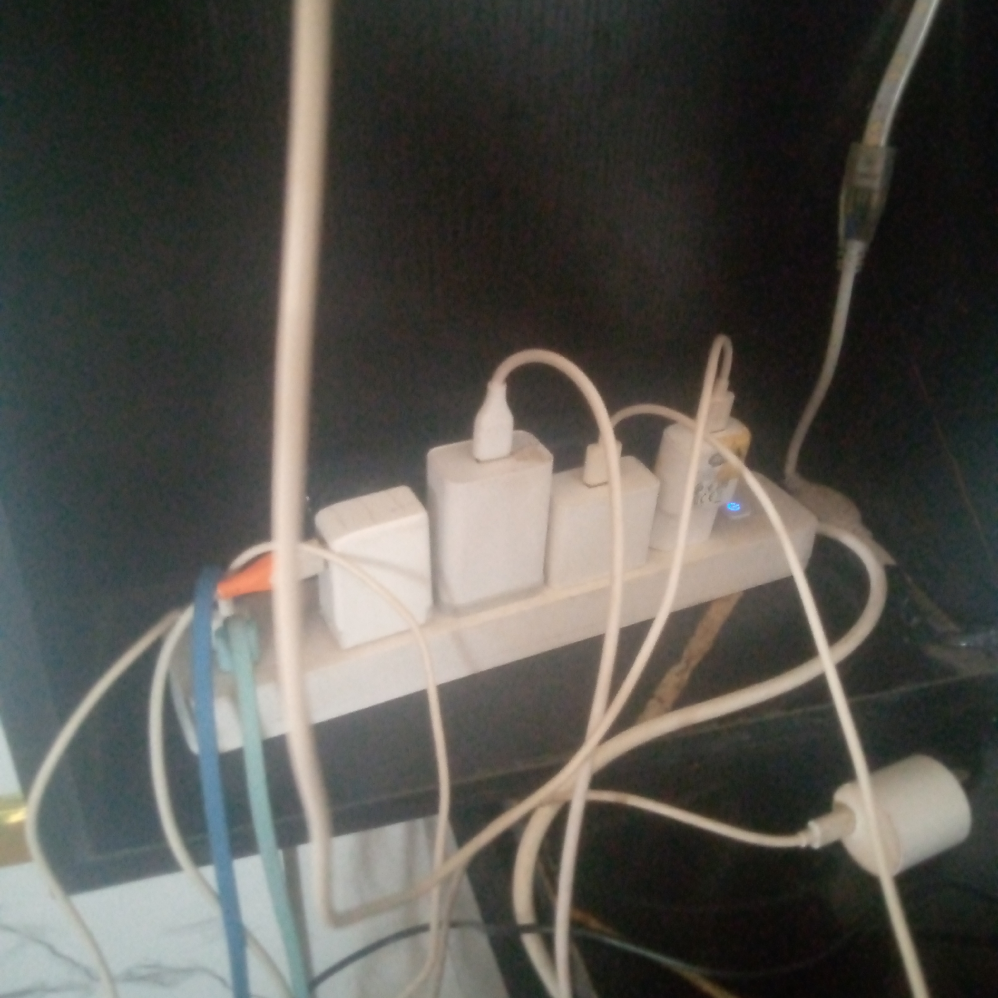 The image displays a white multi-outlet power strip situated horizontally on a dark, featureless surface, likely a shelf or desk, within an indoor setting. Five white rectangular USB wall adapters of varying sizes are plugged into the power strip; two larger adapters are centrally positioned, flanked by three smaller ones. Multiple white USB cables emerge from these adapters, forming a tangled array. A blue LED indicator is illuminated on the second adapter from the right, signifying power or activity. On the far left, two cables with distinct blue and orange insulation/tips are plugged into the power strip. To the upper right, a transparent-jacketed cable, resembling an Ethernet cord with a visible RJ45 connector, is connected and extends downward. Another standalone white barrel-jack power adapter is visible on the far right, connected by a looping white cable. The scene depicts a functional but disorganized charging and power distribution point.