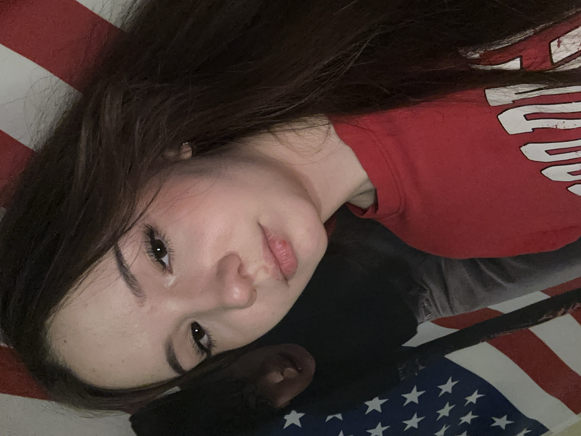 A young woman with long, dark hair is posing for a selfie. She is wearing a red t-shirt. Behind her, an American flag is visible, as well as a blurred figure of another person. The lighting is dim and appears to be artificial, casting shadows on the subjects. The background is cluttered, with the American flag partially obscuring the environment. There are no discernible landmarks or signs that would indicate a specific location in Naberezhnye Chelny, Russia.