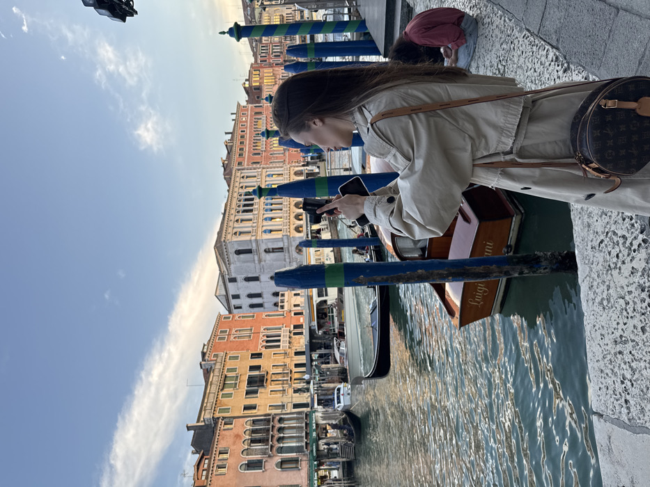 The image depicts a canal scene in Venice, Italy, likely the Grand Canal, under a clear, light blue sky with minimal cloud cover, suggesting daylight. In the foreground right, a woman with long, dark hair, wearing a light-beige trench coat and a circular dark brown crossbody bag with a monogram pattern (Louis Vuitton), stands on a rough-textured grey stone embankment. She holds a black smartphone horizontally with both hands, aiming it towards the canal and the buildings on the far bank, appearing to photograph or video the scene. Behind her, partially visible in the lower right, another individual is seated on the embankment, wearing a reddish-brown long-sleeved garment, with their head lowered. The canal itself displays rippled, teal-colored water. Numerous vertical mooring poles, some painted with blue and green stripes, are visible along the canal's edge. Several boats occupy the water: a dark gondola with an individual propelling it, a small wooden motorboat named "Luigi Rian" moored to a pole, and a white motorboat further in the distance. The background consists of a continuous line of historic, multi-story buildings, varying in color from pink and orange to yellow and white, featuring numerous windows, balconies, and classical architectural elements.