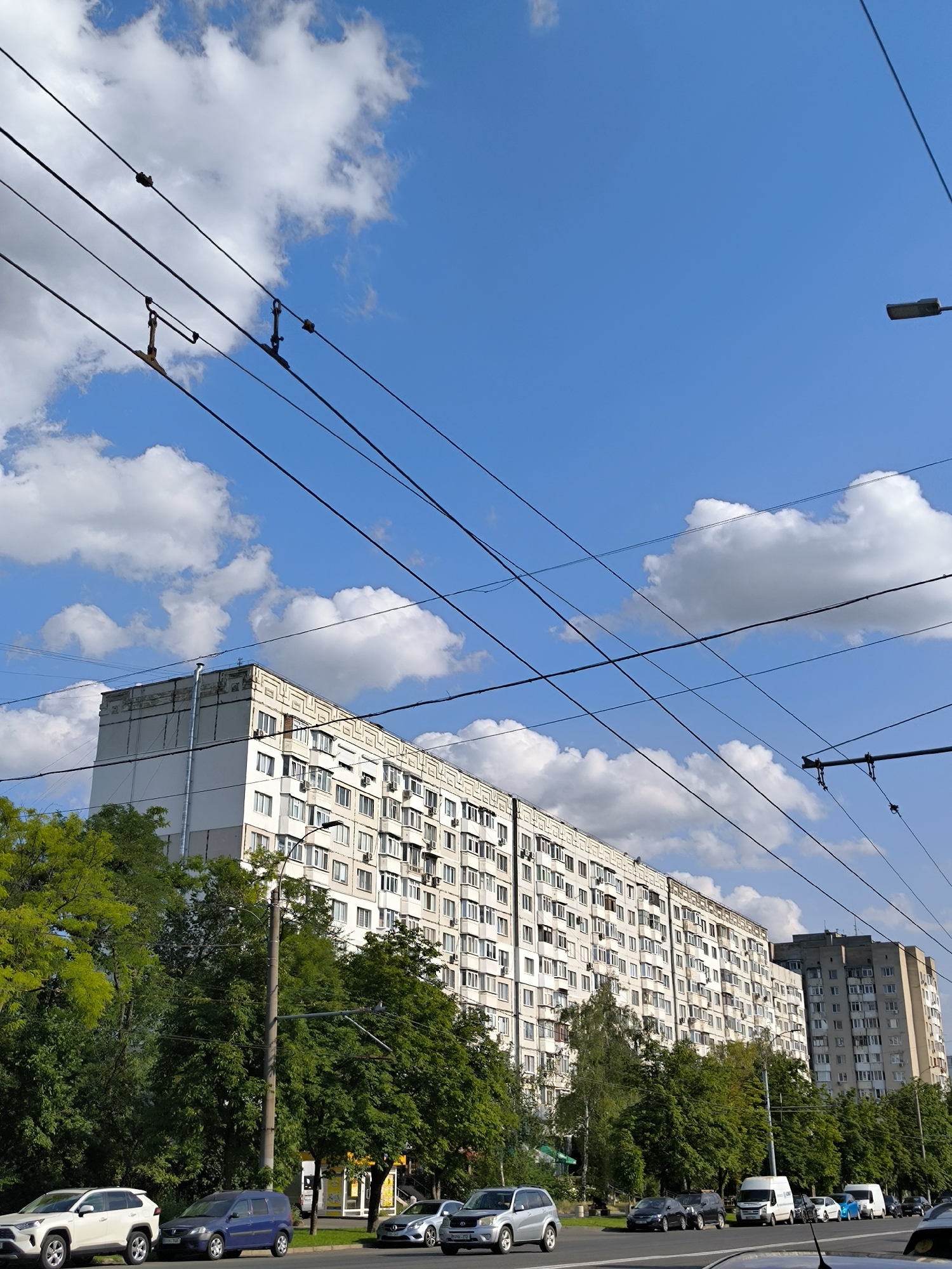 The image depicts a street scene in Chișinău, Moldova, under a bright blue sky dotted with white cumulus clouds. Overhead, a complex network of electrical wires crisscrosses the frame, likely for trolleybuses or trams, indicated by the insulators and attachment points visible.
In the background, two large, multi-story apartment buildings dominate the skyline. The one on the left is a long, cream-colored block with numerous windows, balconies, and air conditioning units, suggesting a residential area. It has a distinctive decorative frieze near the roofline and a large metal pipe running vertically along one side. The building to its right appears to be slightly shorter and clad in gray concrete.
A row of mature green trees lines the street between the buildings and the road, providing a lush natural element. Several cars are parked or driving on the road, including a white SUV, a dark blue minivan, a silver sedan, and a white van. A street lamp is visible on a pole amidst the trees. The overall impression is an urban environment with a mix of residential architecture and greenery.