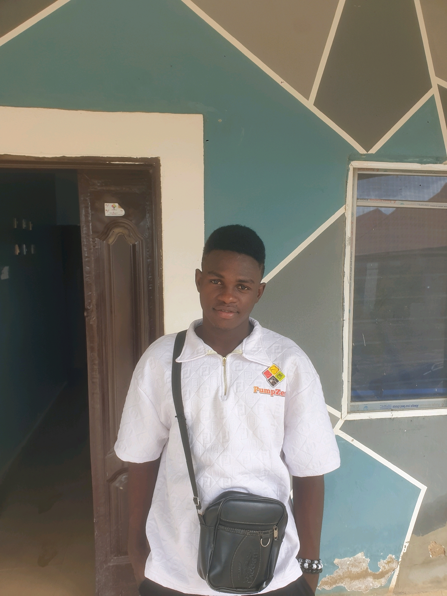 A young Black male stands in front of a brown door and a wall with a geometric pattern. The male is wearing a white, textured polo shirt with a zipper and a logo that reads "PumpZe." He also has a black crossbody bag and two beaded bracelets on his left wrist. The background consists of a building with a teal and gray geometric design on the exterior wall. To the right of the male is a window with a blue sky reflected in it. The location appears to be Mangu Town, Nigeria, indicated by the context provided.