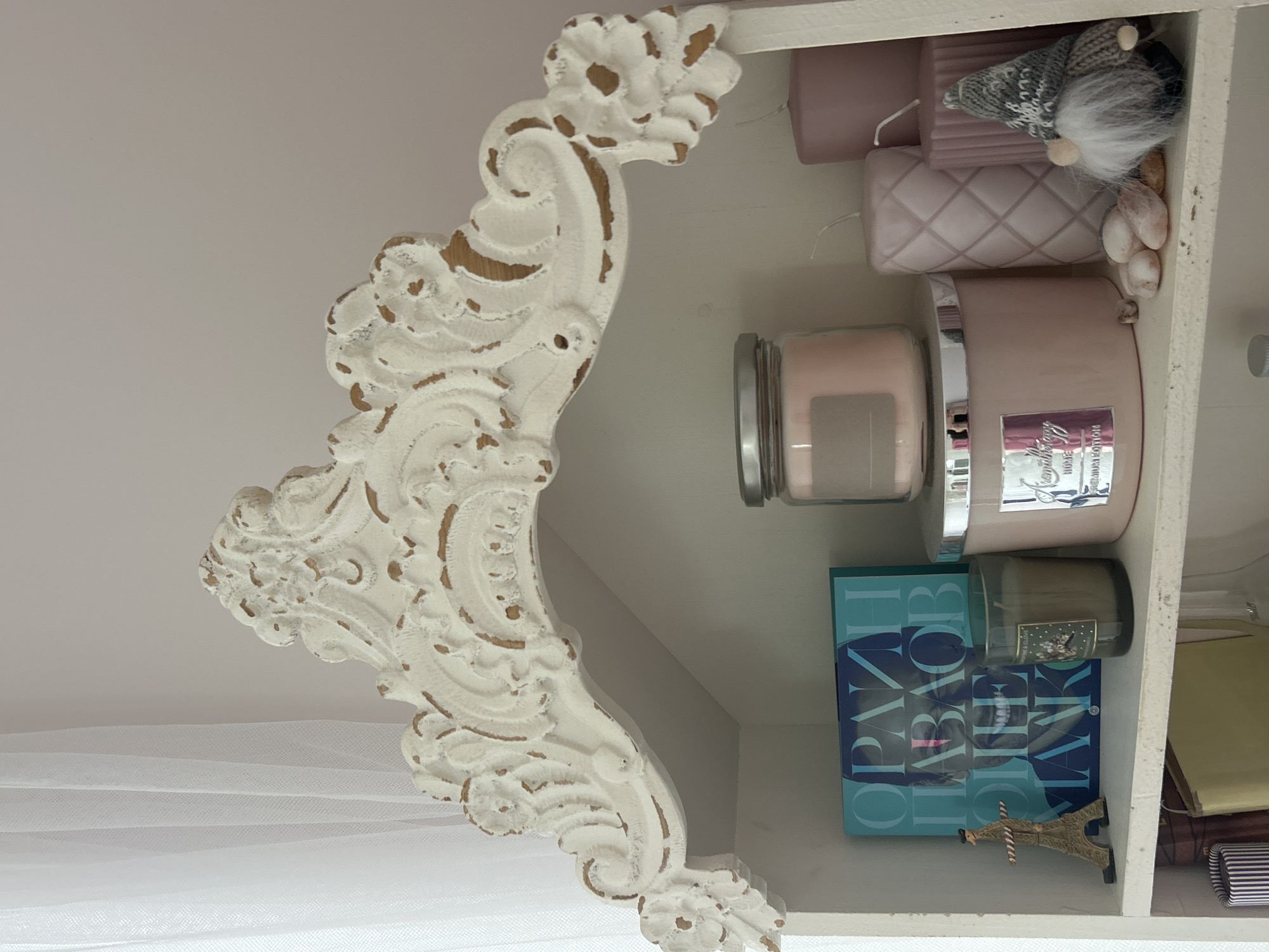 The image depicts a corner of a shelf, presumably inside a home or shop, not specifically indicating a location in Plovdiv, Bulgaria. The shelf is white, and features an ornate, distressed white decorative bracket on the left side. On the shelf, there are several items: two cylindrical candles in light pink, a small gnome-like doll with a grey hat and white beard, several small white shells, a large jar candle with a silver lid and a light pink wax, a smaller green candle, a teal-colored book with Cyrillic script, a small Eiffel Tower replica, and some notebooks or papers. The background is predominantly white. A sheer white fabric is visible in the bottom left corner.