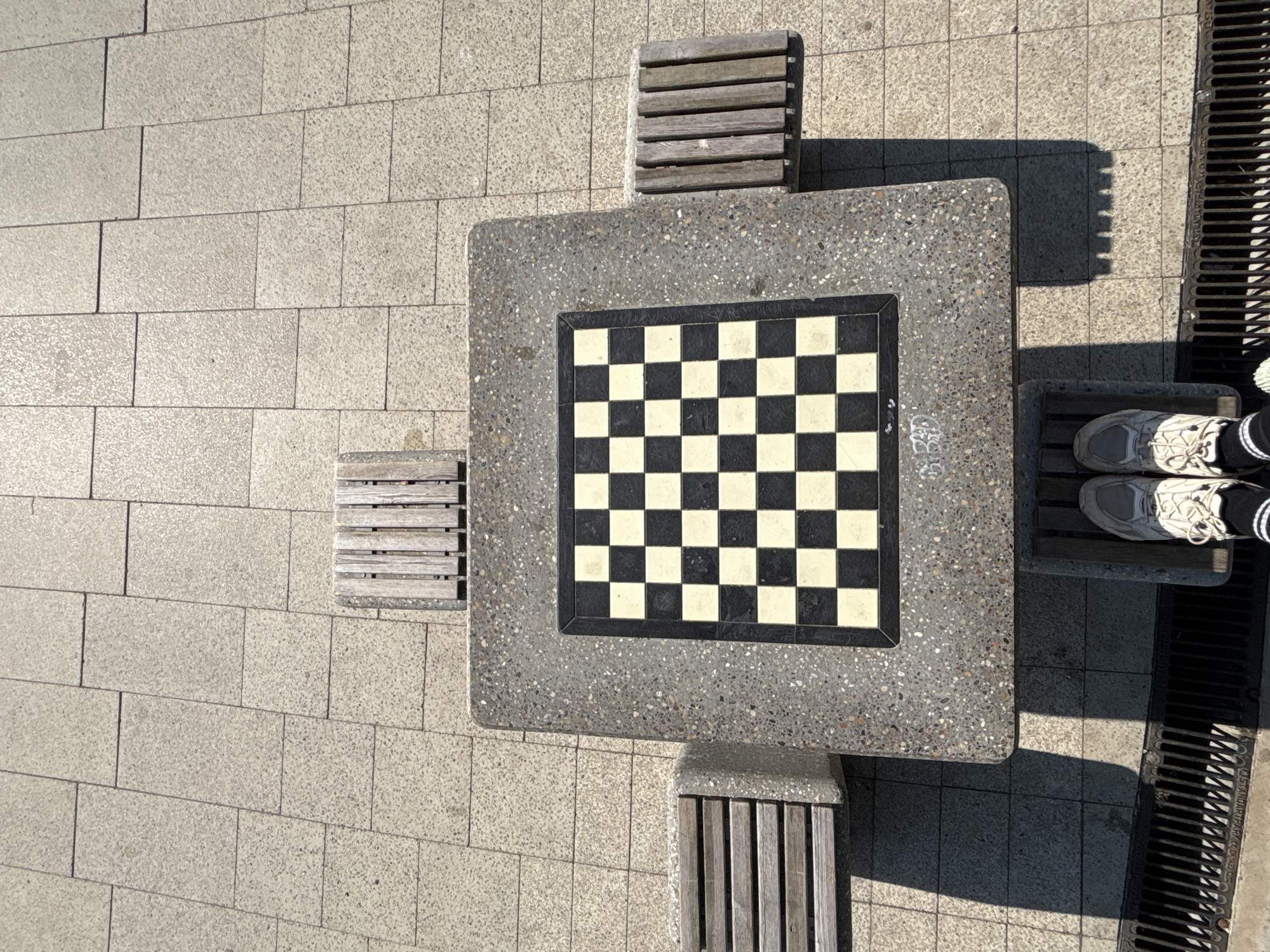 A top-down view shows a built-in chessboard table and benches outdoors. The table has a gray, speckled concrete surface with a black and white checkered chessboard inlaid in the center. Attached to each of the table's four sides is a small wooden bench with multiple slats. The table sits on a paved area constructed of light gray rectangular blocks. To the right of the scene is a metal grate covering a drain. The feet of a person wearing athletic shoes and striped socks are visible standing on one of the benches near the grate. The light indicates a sunny day, casting shadows from the benches and table. Location cannot be definitively determined.