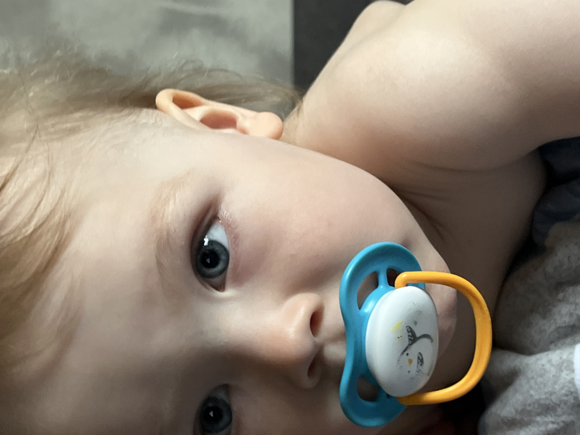 A close-up, slightly angled, indoor shot features a baby lying on its side with a blue and yellow pacifier in its mouth. The baby has fair skin, light blond hair, and blue eyes. The right ear is visible, and the baby's face fills the majority of the frame. Behind the baby is a blurry background of a dark gray surface and a grayish-white textured material. There is no evidence to suggest the location of the photo or any indication it was taken in the city of Chaikovsky, Russia.