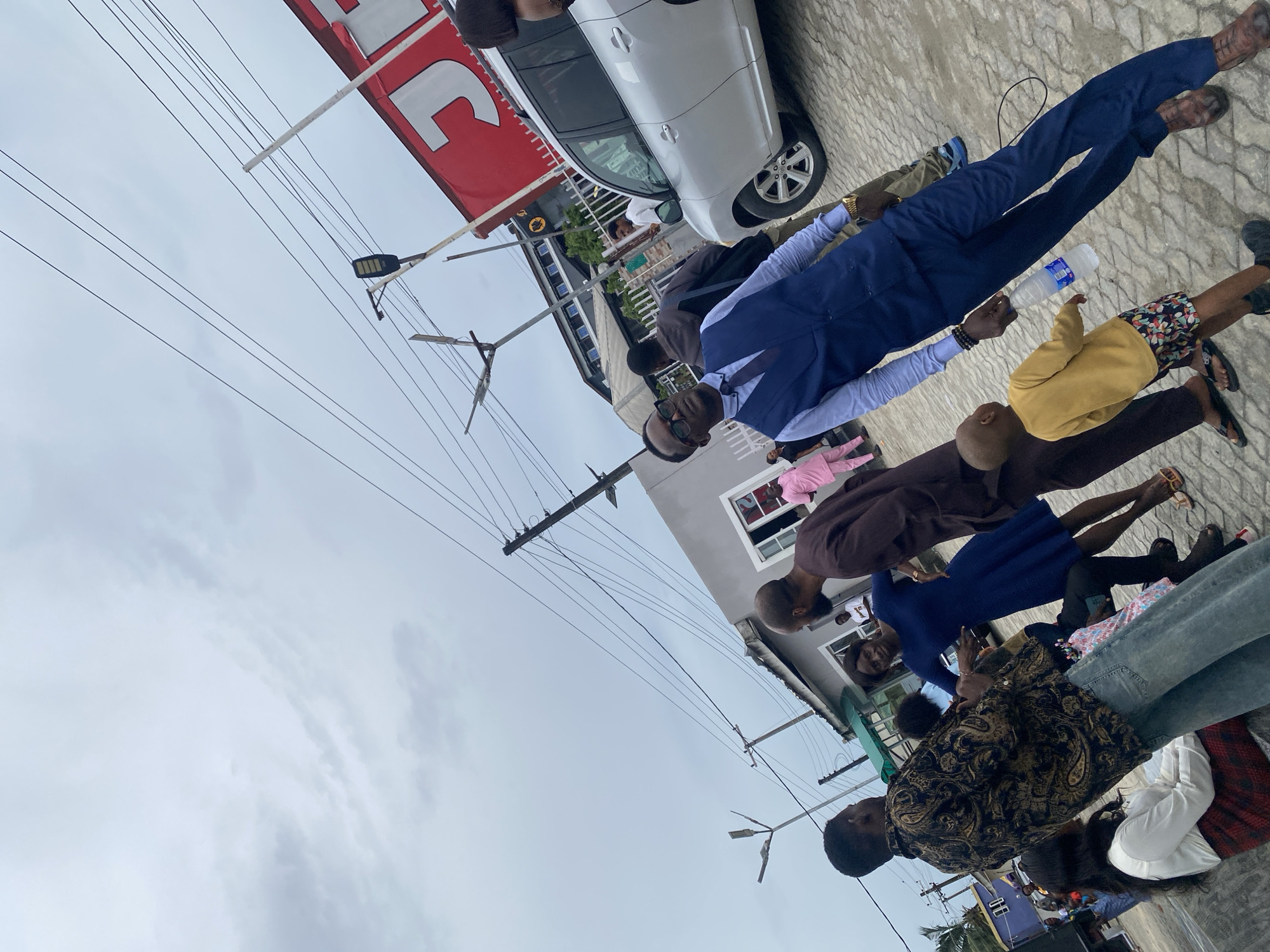 The image appears to be taken outdoors in a commercial or residential area. Several people are gathered, seemingly observing or participating in an event. They are diverse in age and are dressed in a range of casual and formal attire. One man is wearing a suit and holding a water bottle. Others are in dresses, jeans, and other informal wear.
In the background, there is a white car parked near a building with a red sign, possibly a storefront. Overhead power lines crisscross the sky, and the sky itself appears cloudy. Buildings are visible, suggesting an urban environment.
Given the mention of Lekki, Nigeria, it is presumed that this image was taken in Lekki. This observation is based on the metadata provided rather than visual cues within the image. Further details or specific location information would require a more detailed analysis.
