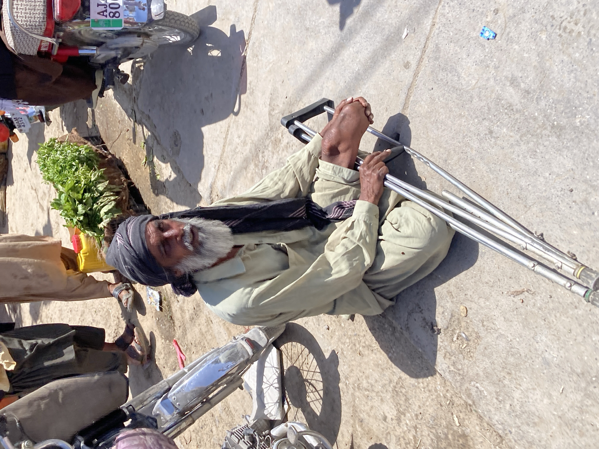 An elderly man with a gray beard and a head covering is sitting on the ground with his legs crossed. He has a pair of crutches in his hands. The setting appears to be an outdoor area of Quetta City Tehsil, Pakistan. There is a motorcycle parked nearby, and what looks like greens for sale. Several people are standing nearby. The ground appears to be concrete.