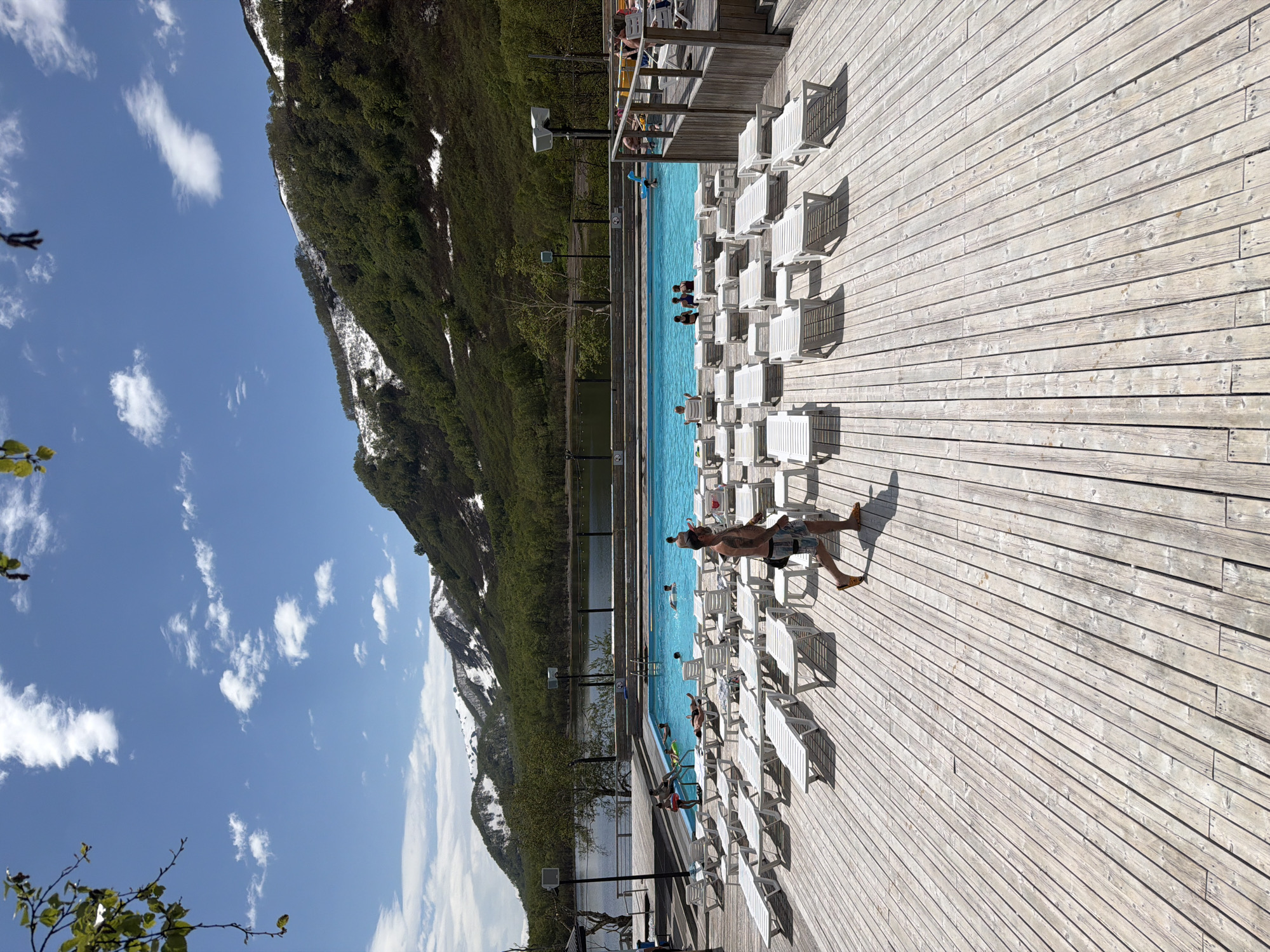 In Paratunka, Russia, a man with tattoos on his arms and legs, wearing patterned swim shorts and yellow sandals, walks across a weathered wooden deck. The deck is lined with rows of empty white lounge chairs, some with umbrellas. In the background, a bright blue swimming pool is filled with people swimming and relaxing. Beyond the pool, a dark lake is visible, bordered by a tree-lined shore. Majestic, snow-capped mountains rise in the distance under a clear blue sky dotted with white clouds. The scene appears to be a resort or recreational area, taking advantage of the natural landscape.