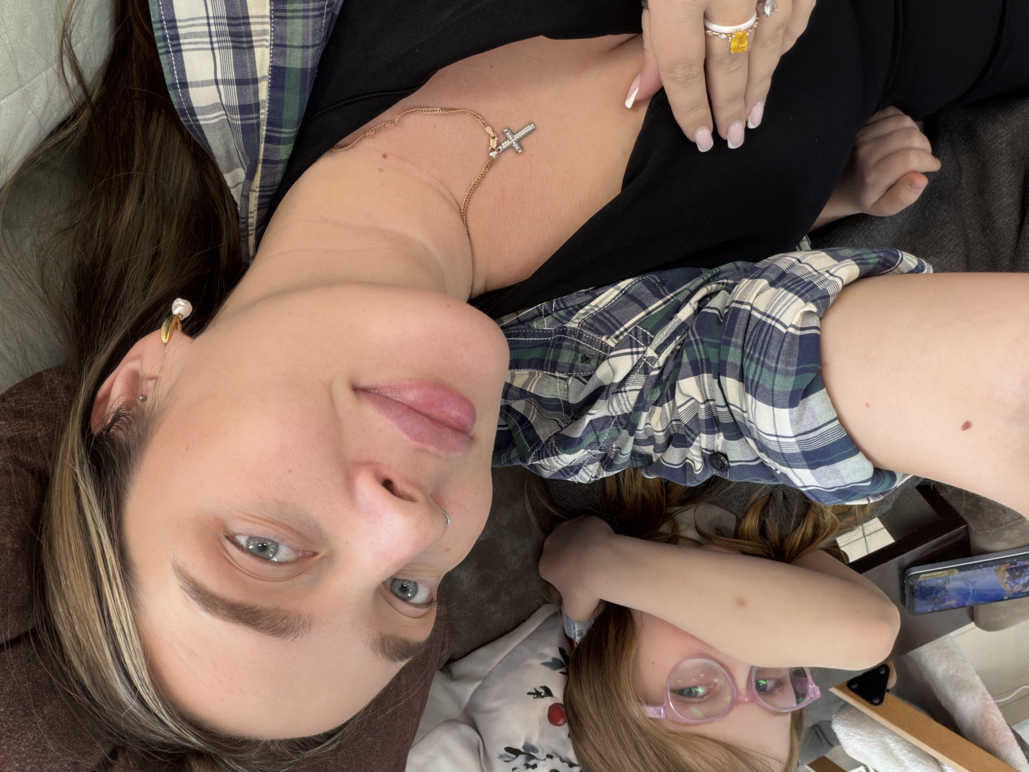 An overhead, sideways-oriented image captures a woman and a child lounging indoors, likely on a sofa or bed.
In the foreground, an adult woman lies on her back, looking directly at the camera with a calm expression. She has long, dark hair with blonde highlights, clear blue eyes, and a small silver nose ring. She wears a golden ear cuff with a white pearl on her right earlobe. Around her neck is a delicate gold chain featuring a small, ornate cross pendant. Her attire consists of a dark blue and green plaid button-up shirt worn over a black top. Her right hand, adorned with a white and yellow ring and another clear-stoned ring, rests near her shoulder, showcasing neatly manicured nails with a French tip style.
Partially visible in the background, to the right, is a child with light brown hair. The child wears pink-framed glasses and a light-colored top, possibly decorated with a festive red berry design. Their right arm is bent, partially obscuring their face, and they appear to be looking at a mobile phone or tablet, a portion of which is visible with content on its screen.
The setting appears to be a cozy indoor environment, with brown upholstered furniture or bedding providing the background for the woman. The overall impression is one of relaxed downtime, possibly a casual moment captured as a selfie or by a third party. No visible text is present within the image. The location context provided is Chaikovsky, Russia.
