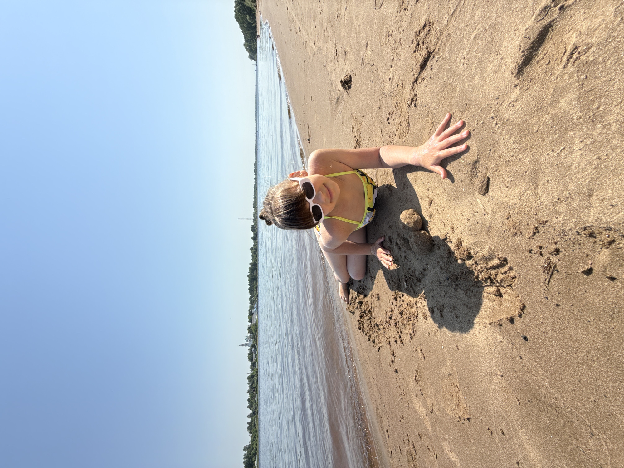 A child wearing a yellow bathing suit and white sunglasses kneels on a sandy beach in Шебунино, Russia. The child faces the camera, with their right hand outstretched on the sand. The beach stretches along the edge of a body of water, with trees lining the opposite shore. The sky is clear and blue.