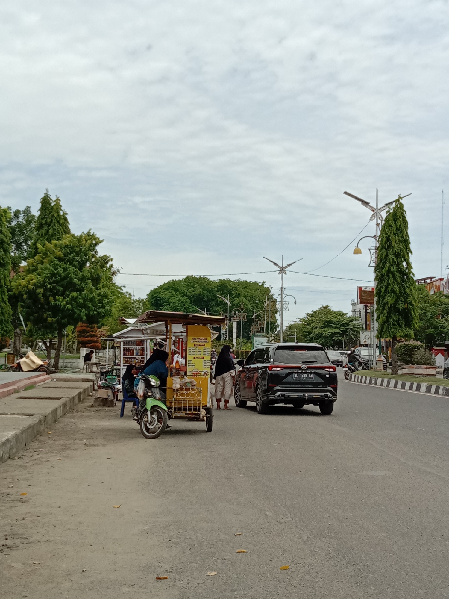 The image captures a street scene in Lhokseumawe, Indonesia. A black Toyota Avanza with license plate BL 1898 KB is visible on the right side of the street, facing away from the viewer. Two people stand near a food vendor stall pulled by a motorcycle. The vendor stall is yellow and has various food items displayed. The individual near the stall appears to be engaging with the vendor. The street is lined with trees, and there are decorative street lights. Other vehicles are visible further down the street. The sky is overcast.
