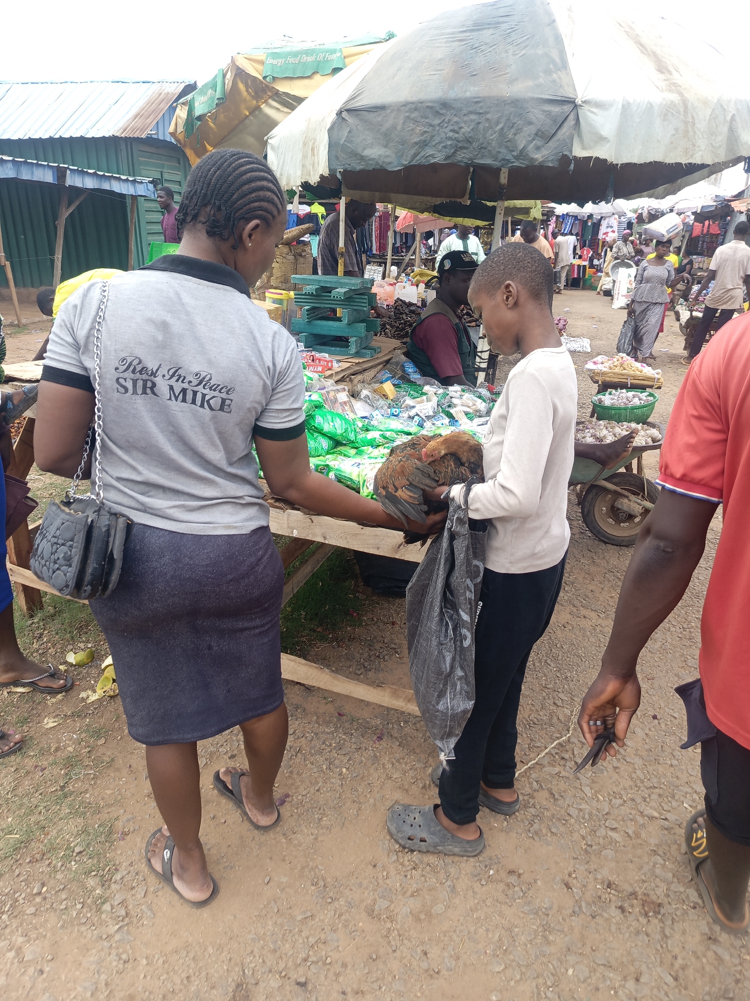 The image depicts a bustling market scene in Akwanga, Nigeria. A woman with braided hair is facing a younger person who is holding a chicken in a plastic bag. They are both standing at a wooden table filled with goods. The woman is wearing a gray t-shirt with "Rest In Peace SIR MIKE" printed on the back, a dark skirt, and sandals, and she has a black purse hanging from her shoulder. The younger person is wearing a light-colored long-sleeve shirt, dark pants, and gray clogs.
A man in a red shirt is partially visible on the right side of the frame. In the background, there are other people and market stalls beneath an umbrella. A wheelbarrow filled with goods and other market items are also visible. The ground is dirt and gravel.