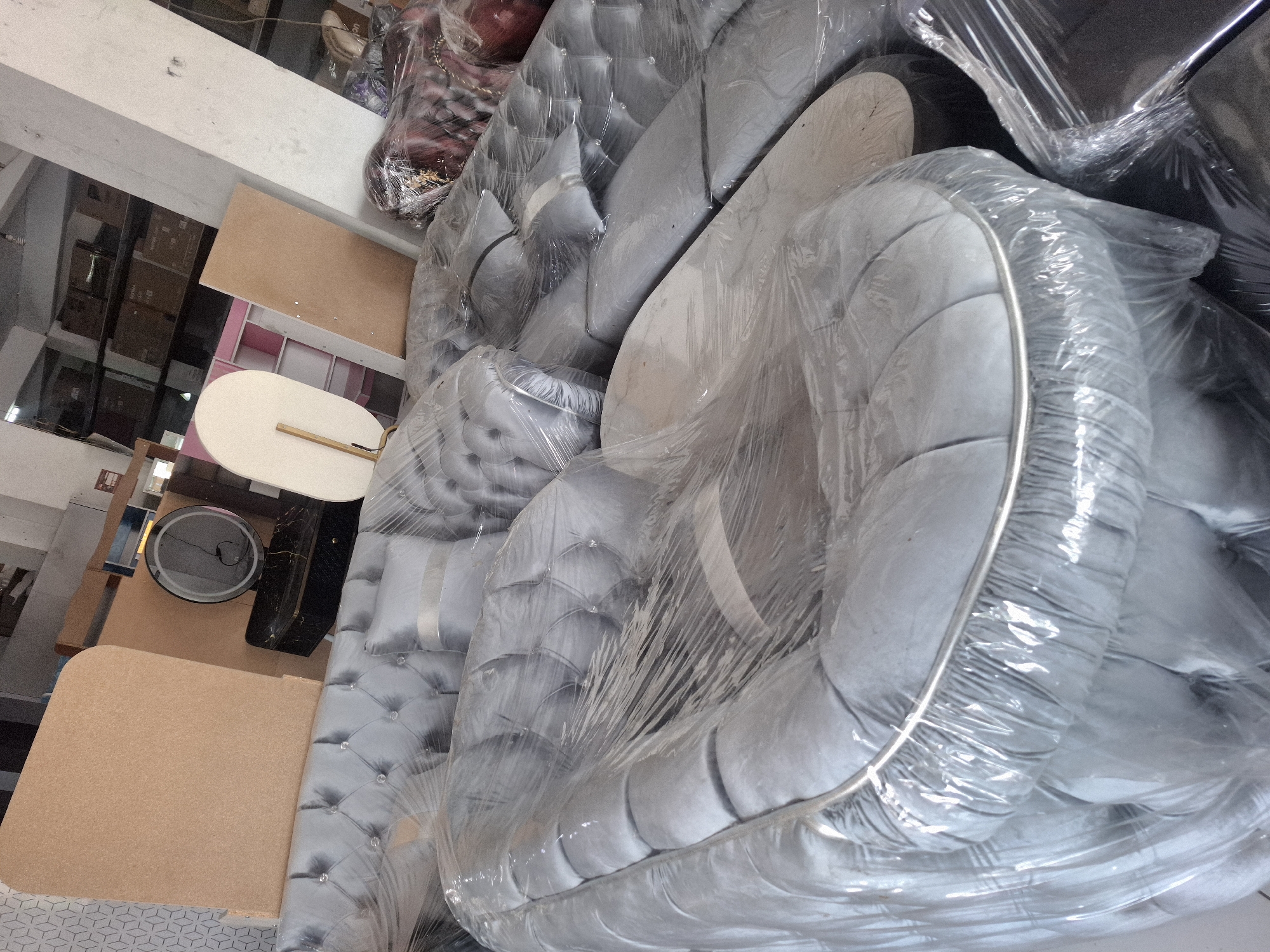 In Banjarmadu, Indonesia, a furniture store displays a collection of upholstered furniture, primarily sofas, wrapped in clear plastic. The scene is dimly lit, with the light source appearing to be overhead fluorescent lights or natural light coming from windows or an open doorway. The furniture is predominantly in shades of gray, with some pieces featuring tufted detailing and decorative crystal accents. In the background, there are other furniture items, including a dresser with a round mirror and some shelving units. Boxes and other miscellaneous items are stacked in the background, suggesting a retail or storage environment. The overall impression is that of a showroom or warehouse filled with new furniture, ready for sale or distribution.