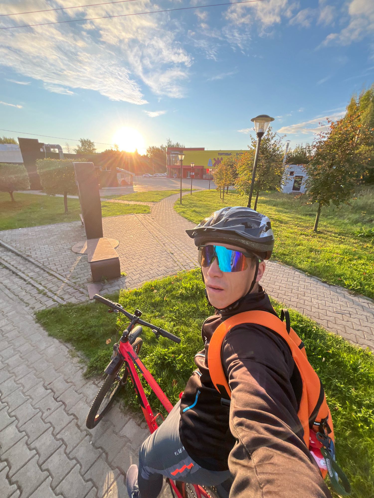 A selfie taken by a person wearing a black helmet with a white and grey design and reflective blue and orange cycling glasses, with an orange backpack, is shown. The person is sitting on a red bicycle with black handlebars and a black seat. They are wearing a black jacket and grey cycling shorts. The bicycle is parked on a paved walkway next to a grassy area. In the background, a modern sculpture is visible, along with trees, buildings, and a street with cars. The sun is setting, casting a warm glow on the scene. The location is likely a park or public space in the city of Chaikovsky, Russia, given the style of architecture and the presence of a sculpture.