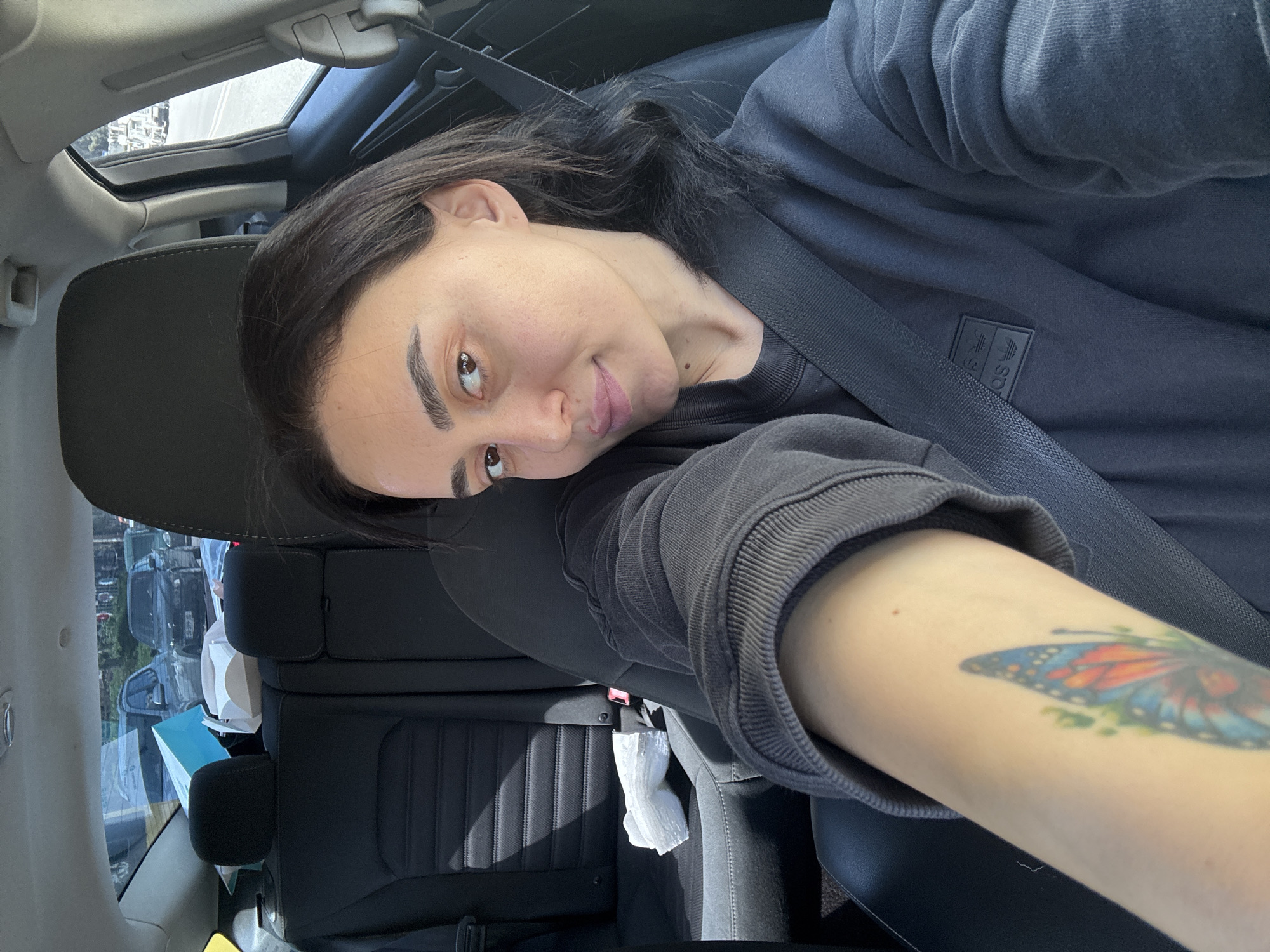 A young woman is sitting in the driver's seat of a car, wearing a dark grey Adidas sweatshirt and a seatbelt. She has dark hair and is looking at the camera with a slight smile. Her left arm is extended towards the camera, displaying a butterfly tattoo. The interior of the car is visible, including the dashboard and back seats. Through the sunroof and side window, cars are parked in front of what appear to be buildings, suggesting a parking area or street in a city. Based on general architectural styles, the image was likely taken in Tbilisi, Georgia.