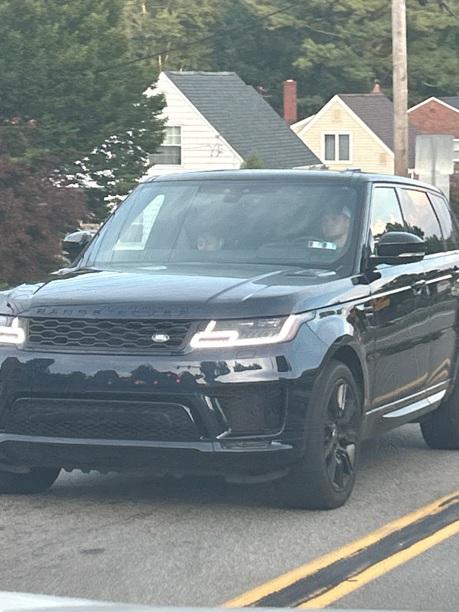 A dark blue Range Rover is driving on a paved road with yellow double lines in the foreground. The vehicle's headlights are on. Inside the vehicle, a person with fair skin and dark hair is visible in the driver's seat. Another person with lighter skin and dark hair is in the passenger seat. Behind the Range Rover, a suburban neighborhood is visible, with houses and trees. The architecture suggests a residential area in the United States, potentially the city of Pittsburgh, USA, based on the style of houses and street markings.