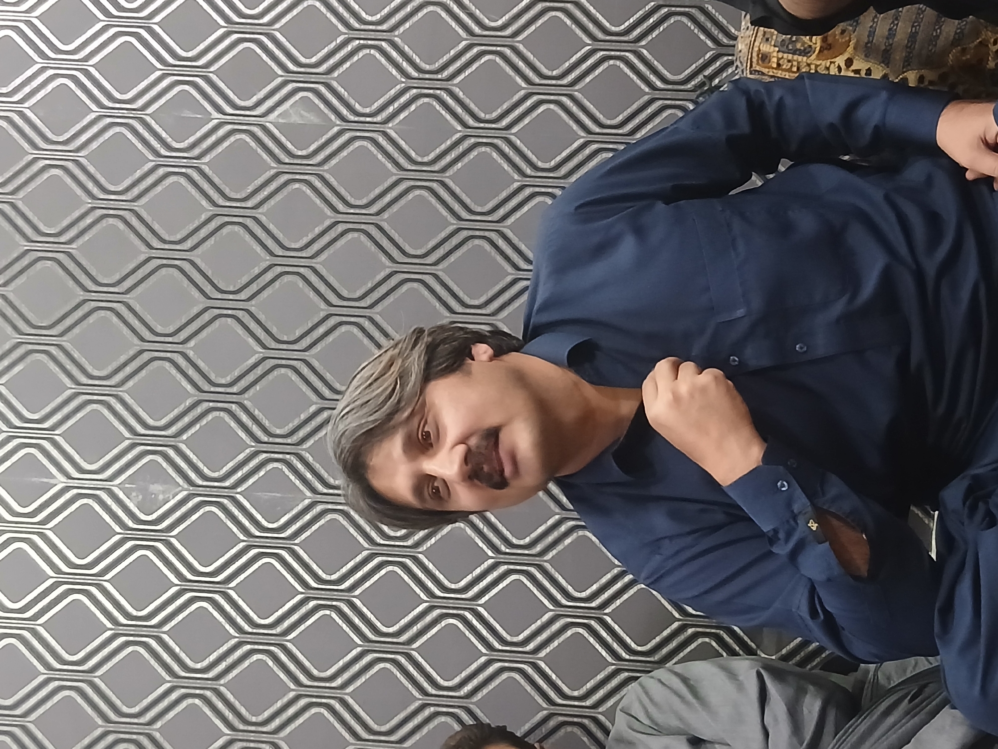 A man wearing a navy blue button-down shirt is seated against a patterned grey and white wall, possibly indoors in Quetta City Tehsil, Pakistan. He has dark hair with grey highlights and a mustache. He is looking towards the camera with his hand near his chest. Another person is partially visible in the lower portion of the image, wearing grey clothing. A patterned fabric is also partially visible.
