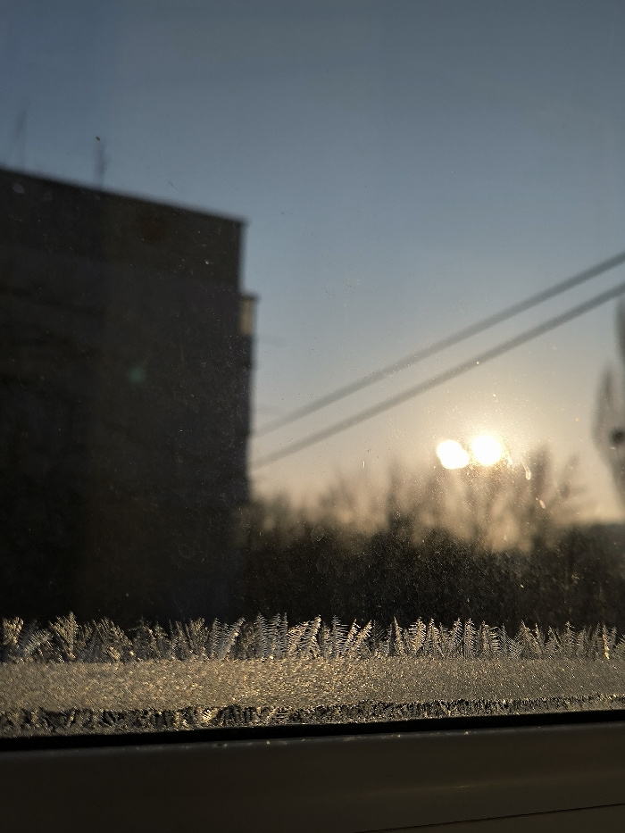 A frosty window obscures a view of a blurry building, power lines, and a hazy sunset. The ice crystals, densely packed at the bottom of the glass, refract the light, creating a shimmering effect. The window's surface shows imperfections and smears, further distorting the scene beyond. The sky transitions from a pale blue to a warm orange near the horizon where the sun's glow is visible.