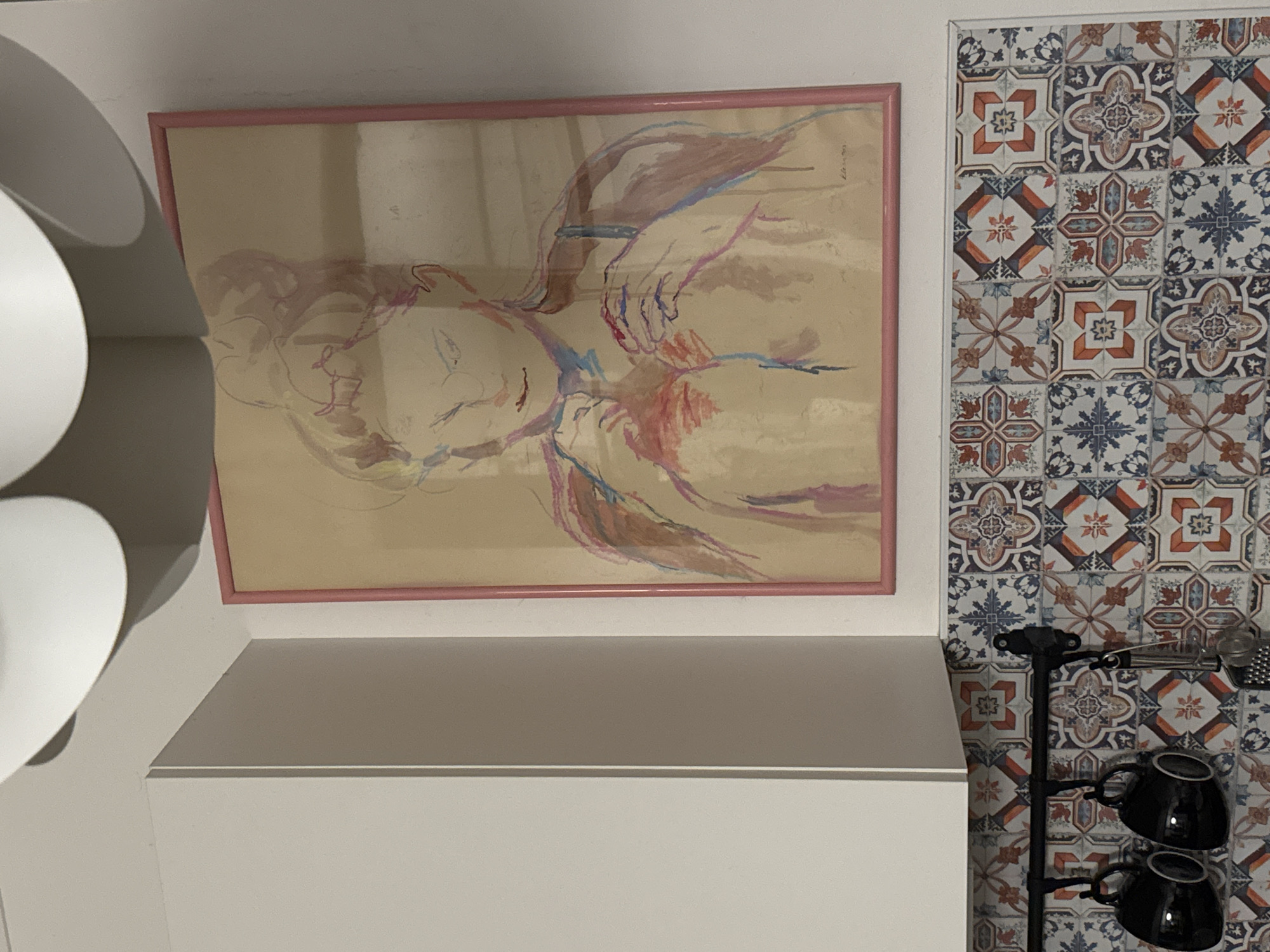 A framed artwork of a woman's upper body, rendered in a loose, abstract style with pastel colors, hangs on a white wall above a section of decorative tile. Two black mugs are suspended from a rack below the artwork. The artwork's frame is a light pink. To the left of the artwork is a portion of a white cabinet. The decorative tiles feature a mosaic pattern of geometric shapes and floral motifs in shades of blue, white, orange, and red.