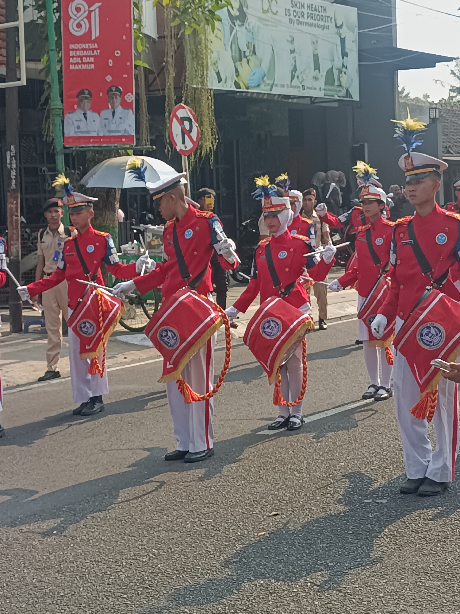 The image depicts a marching band procession in Sleman, Indonesia. In the foreground, several individuals in matching red band uniforms are seen marching. They are wearing white pants, white gloves, and distinctive hats adorned with yellow and blue feathers. Each person carries a drum, which is red with a circular emblem and decorated with orange tassels and braided cords. Some individuals are holding drumsticks, suggesting they are playing the drums. In the middle ground, other participants and onlookers are visible. Behind them, a banner with "81" prominently displayed and text in Indonesian is hung vertically from a pole. Another banner with English text "SKIN HEALTH IS OUR PRIORITY By Dermatologist" is visible in the background. The scene takes place on an asphalt road, with shadows cast by the participants and surrounding trees. The environment suggests an outdoor public event, possibly a parade or celebration.