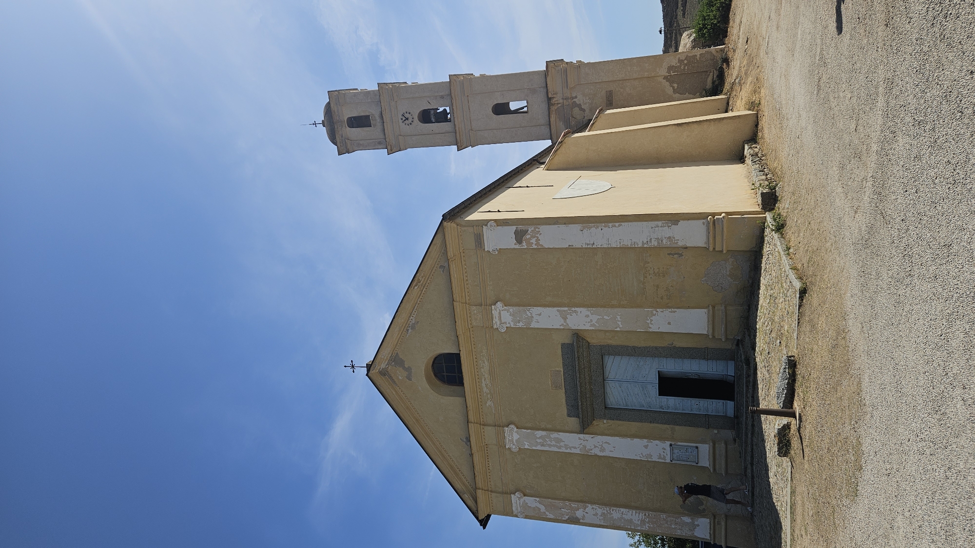 A low-angle, full outdoor shot captures a yellow church with a bell tower in Sant'Antonino, France. The church has a light blue door and a steeple topped with a cross. A single person stands to the left of the door, appearing to take a photograph. The church is situated on a gravel and dirt path, with a clear blue sky overhead. The bell tower is rectangular and has visible bells inside. The church facade features columns and worn paint.