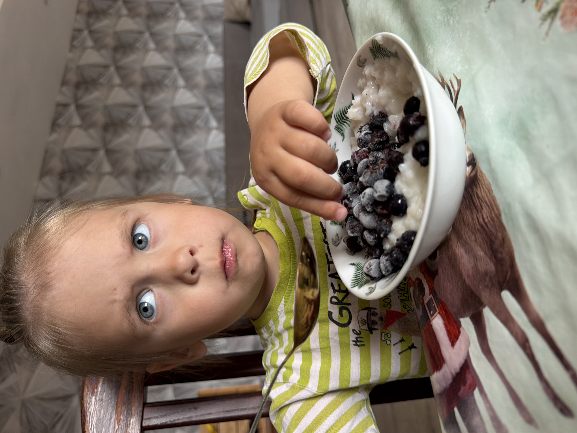 A young child with blonde hair and wide, striking blue eyes is the central subject of this indoor portrait, taken in Chaikovsky, Russia. The image is oriented sideways, with the child's head on the left looking towards the upper right. She is wearing a green and white striped long-sleeved shirt. Her right hand, in the foreground, is holding a white bowl containing a creamy white substance, likely rice porridge, generously topped with dark, frosted berries such as blueberries or blackberries. A golden spoon is partially visible near her chest.
The child's expression is one of curiosity or slight surprise, with her lips slightly parted as she looks directly at the camera. The background features a patterned wall with a light gray, geometric, possibly three-dimensional design. The surface below the bowl is covered by a light-colored tablecloth adorned with a print of a deer and a figure in a red suit resembling Santa Claus. The words "the GREAT" are visible on the child's striped shirt. The lighting suggests an indoor setting, likely during the daytime. The berries appear to be frozen or covered in a light frost.