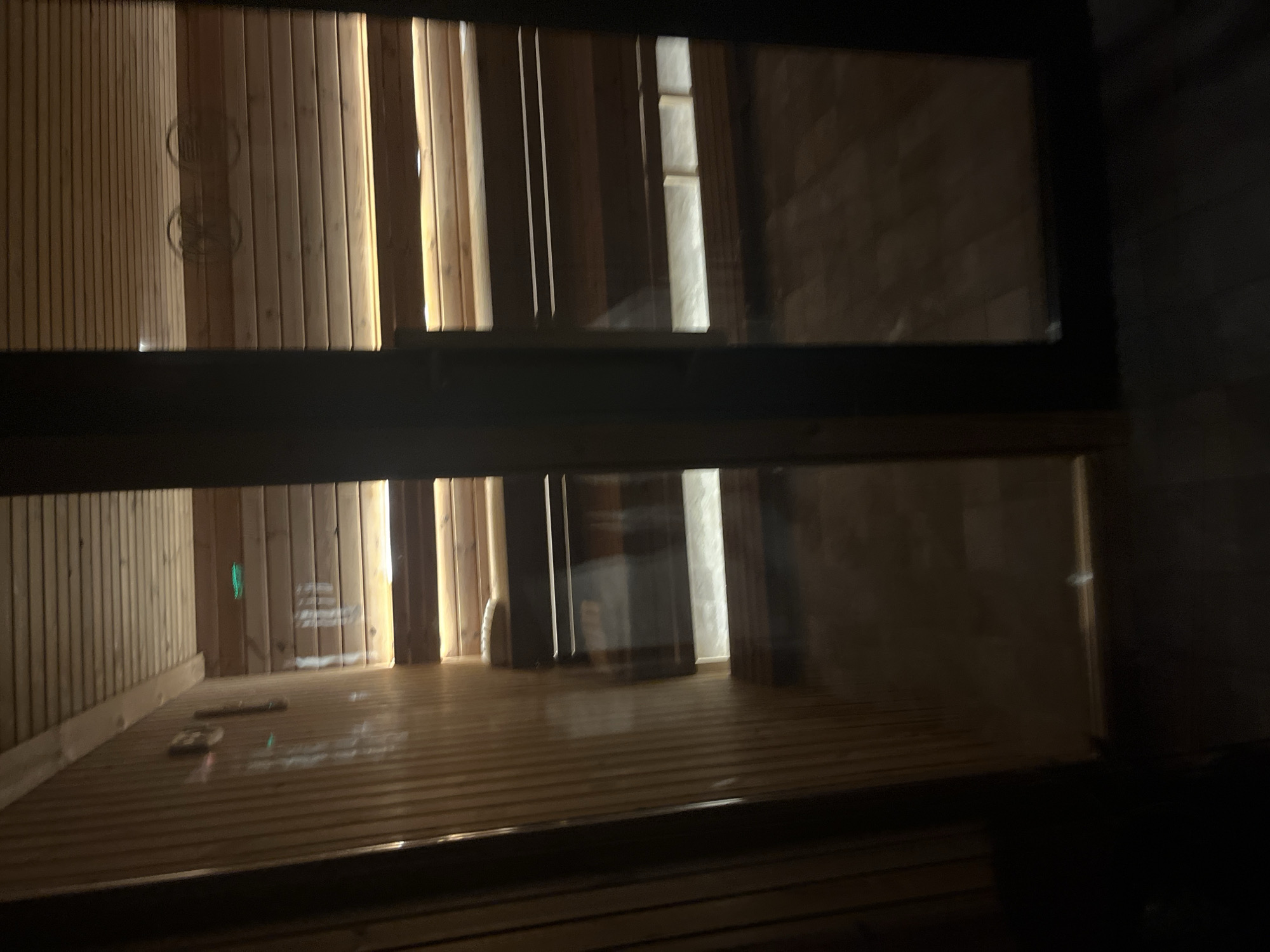 The image, originally rotated 90 degrees counter-clockwise, depicts the dimly lit interior of what appears to be a modern sauna or spa relaxation room. The space is predominantly constructed from light-colored wooden paneling, featuring vertical slats on the walls and horizontal slats on the floor.
Atmospheric, warm lighting emanates from vertical strips hidden behind some of the wooden wall slats, casting distinct bright columns that illuminate the room. A prominent dark, horizontal beam or window frame cuts across the middle of the composition, suggesting a glass partition. Through this partition, reflections of the room's interior are visible.
On the right side, beyond the wooden room and through the glass, a darker, textured wall resembling stacked bricks or stone is faintly visible, indicating an adjacent area or an exterior view. The wooden floor is smooth and reflective, showing slight reflections of the interior lights. Faint green light points or reflections are scattered on the floor and one of the wooden walls. A light-colored, rounded object, possibly a sauna headrest or pillow, rests on the wooden surface in the lower section of the image.
No people are visible in the room. Two faint, circular, stylized patterns or logos are barely discernible on the wooden wall in the upper left, but any embedded text is illegible due to blur and low light. Similarly, some blurry, reflected text is present on the wooden wall in the lower left, but it is unreadable. The overall ambiance suggests a tranquil and serene environment.