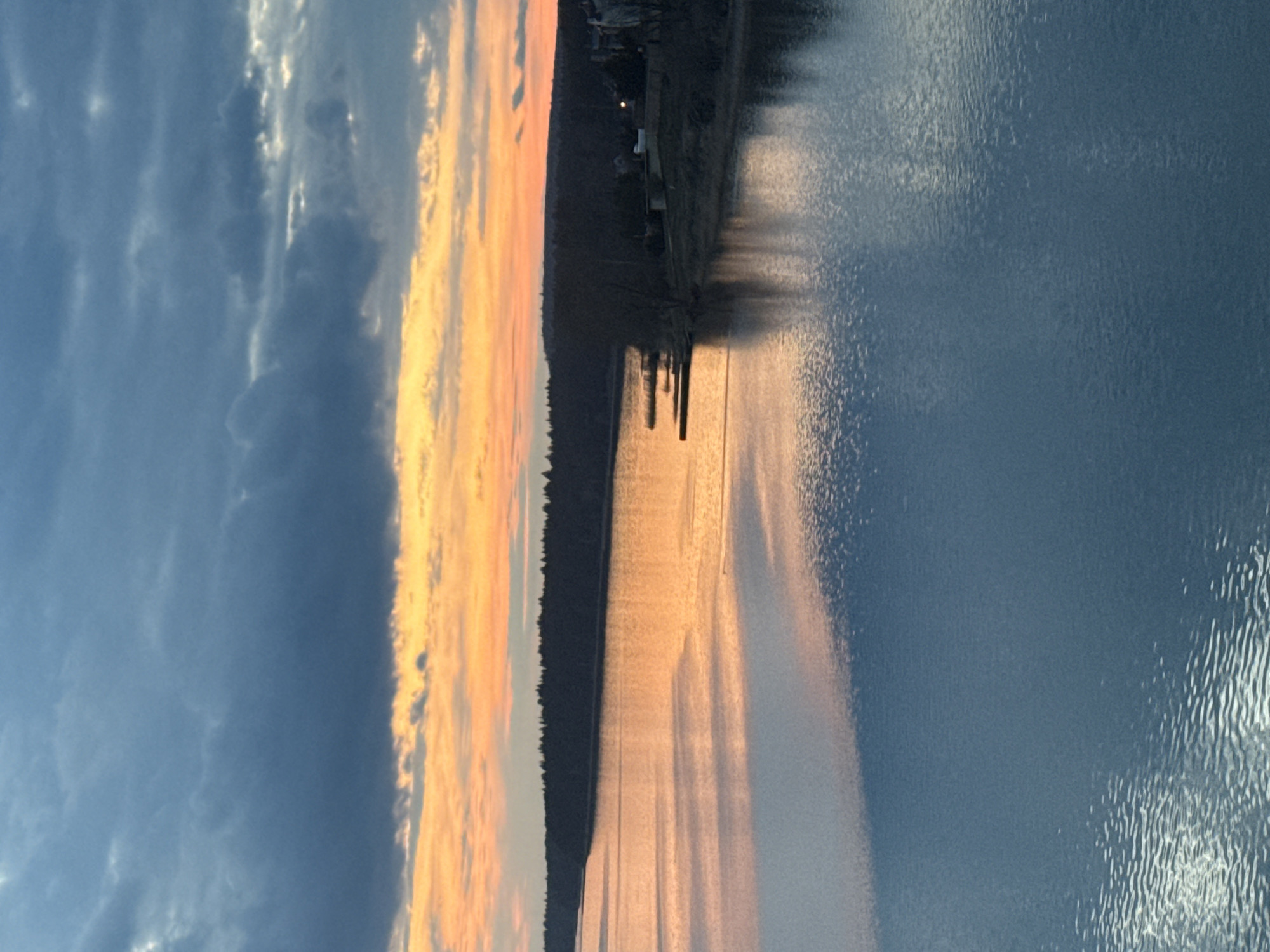 This image captures a tranquil natural landscape during either sunrise or sunset, presenting a broad view of a body of water with a distant shoreline. The sky occupies the upper portion, featuring a striking interplay of dark blue and grey clouds on the left, transitioning to vibrant orange and yellow hues on the right, indicative of the sun's low position.
Along the horizon, a dark, silhouetted treeline or distant landmass stretches across the frame. Barely visible details suggest the presence of a few small structures, possibly houses or buildings, nestled within the landscape. A small set of posts or a rudimentary dock extends into the water from this distant shore, creating faint reflections.
The expansive body of water, likely a lake or large river consistent with the Votkinsk Reservoir in Votkinsk, Russia, dominates the lower half of the scene. Its surface is covered with subtle ripples and textures, beautifully reflecting the sky's colors. The water closer to the horizon glows with the warm, golden light of the sun, while the foreground appears in cooler blue-grey tones, mirroring the cloudier parts of the sky.
The scene evokes a sense of calm and natural beauty during a peaceful twilight hour. There are no visible people or text in the image.