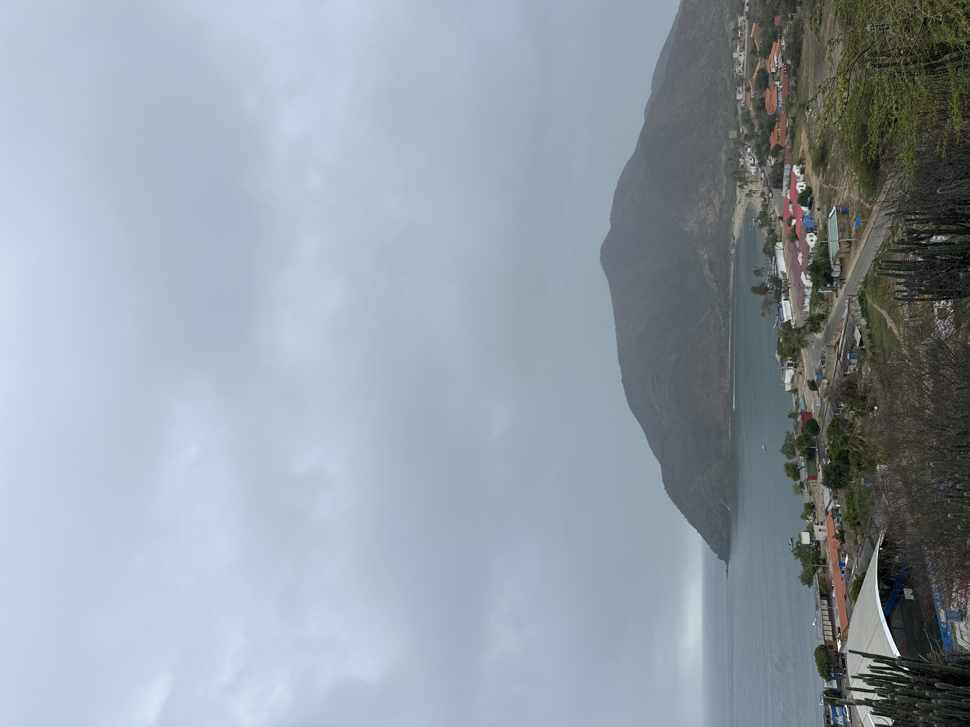 An overcast, elevated view captures a coastal town nestled at the base of a large, rounded mountain. The sea occupies the left third of the frame, with several small boats visible on its surface. Along the shoreline, a collection of buildings with red and orange tile roofs are clustered together, interspersed with smaller structures and vegetation. A road winds through the town, with some vehicles present. In the foreground, cacti and dry brush are prominent. The mountain, a dominant feature, rises from the town and stretches towards the right side of the image, its slopes covered in sparse vegetation. The sky is uniformly grey and cloudy, suggesting a possibly humid or rainy day.