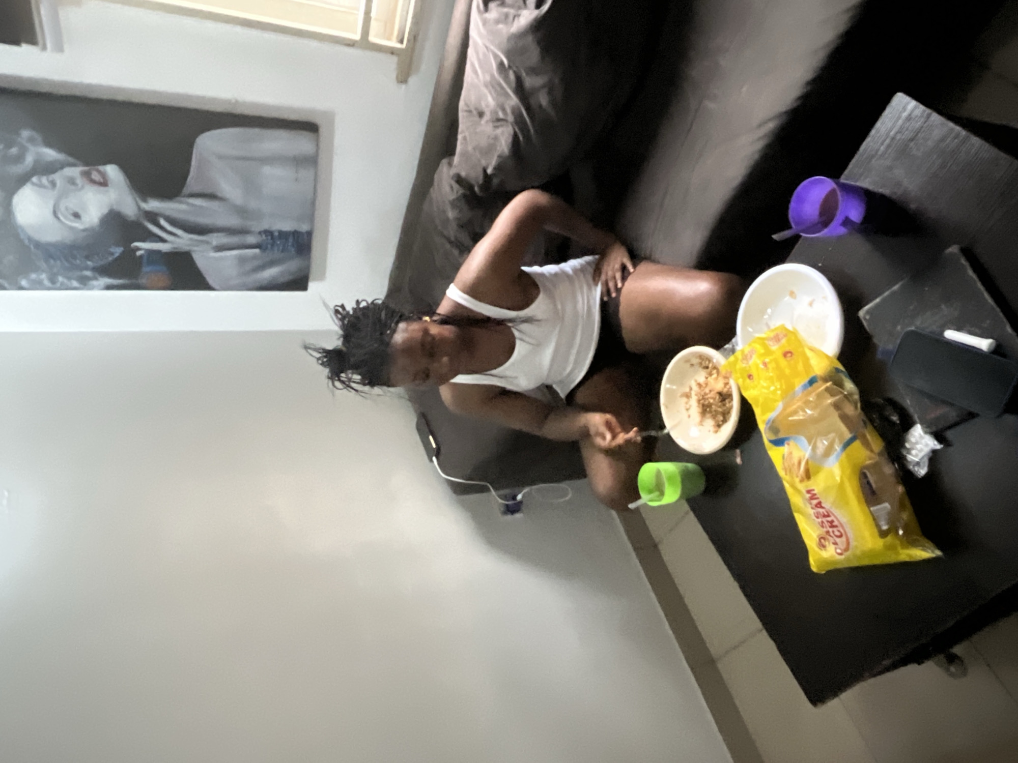 A young Black woman with dark, braided hair pulled up sits on a dark brown couch in an indoor setting. She wears a white tank top and black shorts. In her right hand, she holds a fork, and she is in the process of eating from a white bowl placed on a black coffee table. Beside the bowl, on the table, is a yellow bag of "Classic Cream" biscuits and two cups, one green and one purple, both containing liquid. A phone is also visible on the table. Behind the couch, a framed artwork depicting a stylized portrait of a woman smoking a cigarette hangs on a light-colored wall. The room appears to be a living space, possibly an apartment, located in the city of Abuja, Nigeria.