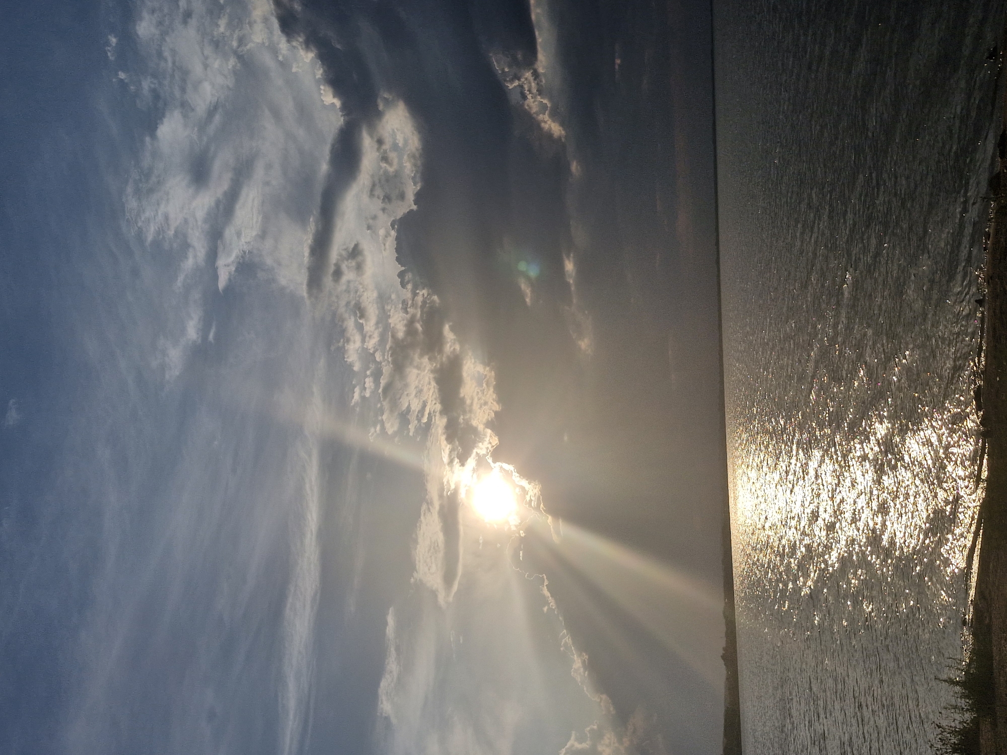 The image depicts a serene natural landscape. A vast body of water, likely a lake, stretches across the foreground. The surface of the water is illuminated by the sun, which is partially obscured by dramatic clouds. Sunbeams pierce through the clouds, creating a shimmering effect on the water's surface. The sky is a blend of blues and grays, with textured clouds contributing to the atmospheric quality of the scene. Along the distant horizon, a faint line of land is visible, suggesting the presence of shores or a coastline. The overall impression is one of natural beauty and tranquility. While the image is a beautiful landscape, it does not contain any specific landmarks or clues that would allow for a precise location within the city of Kokshetau, Kazakhstan.