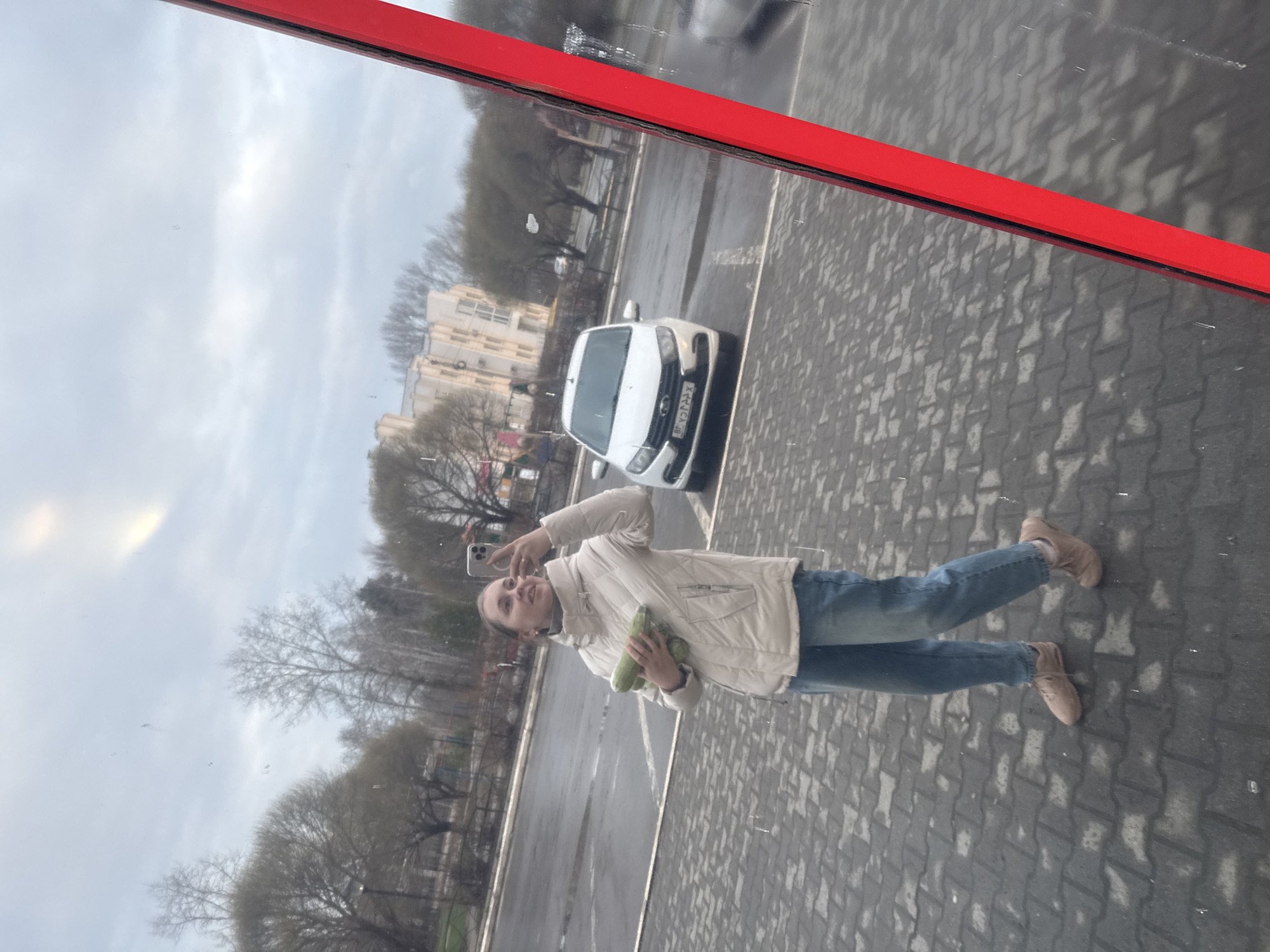 The image displays a full-length reflection of a woman taking a selfie, likely in a large window or reflective glass surface, which is framed by a prominent red border along its top edge. The woman stands upright, holding a smartphone in her right hand to capture the photo, while cradling two long, green vegetables, resembling cucumbers or zucchinis, in her left arm. She is dressed in a light beige or off-white quilted puffer jacket, blue jeans, and light brown flat shoes. A subtle smile is visible on her face.
The reflected environment behind her depicts an outdoor urban setting. The ground beneath her is paved with interlocking gray and dark gray blocks, characteristic of a sidewalk or parking lot, and appears damp in places, suggesting recent rain. In the reflection, a white sedan is parked on the paved area behind the woman. Its license plate features non-Latin characters, specifically Arabic or Persian script and numerals (e.g., ٣٦ و ٧٠٧٠٨). Further in the background, numerous trees with bare branches indicate a late fall, winter, or early spring season. Parts of multi-story buildings can be seen beyond the trees. The sky is overcast with light gray clouds, indicating daytime under cloudy weather conditions.