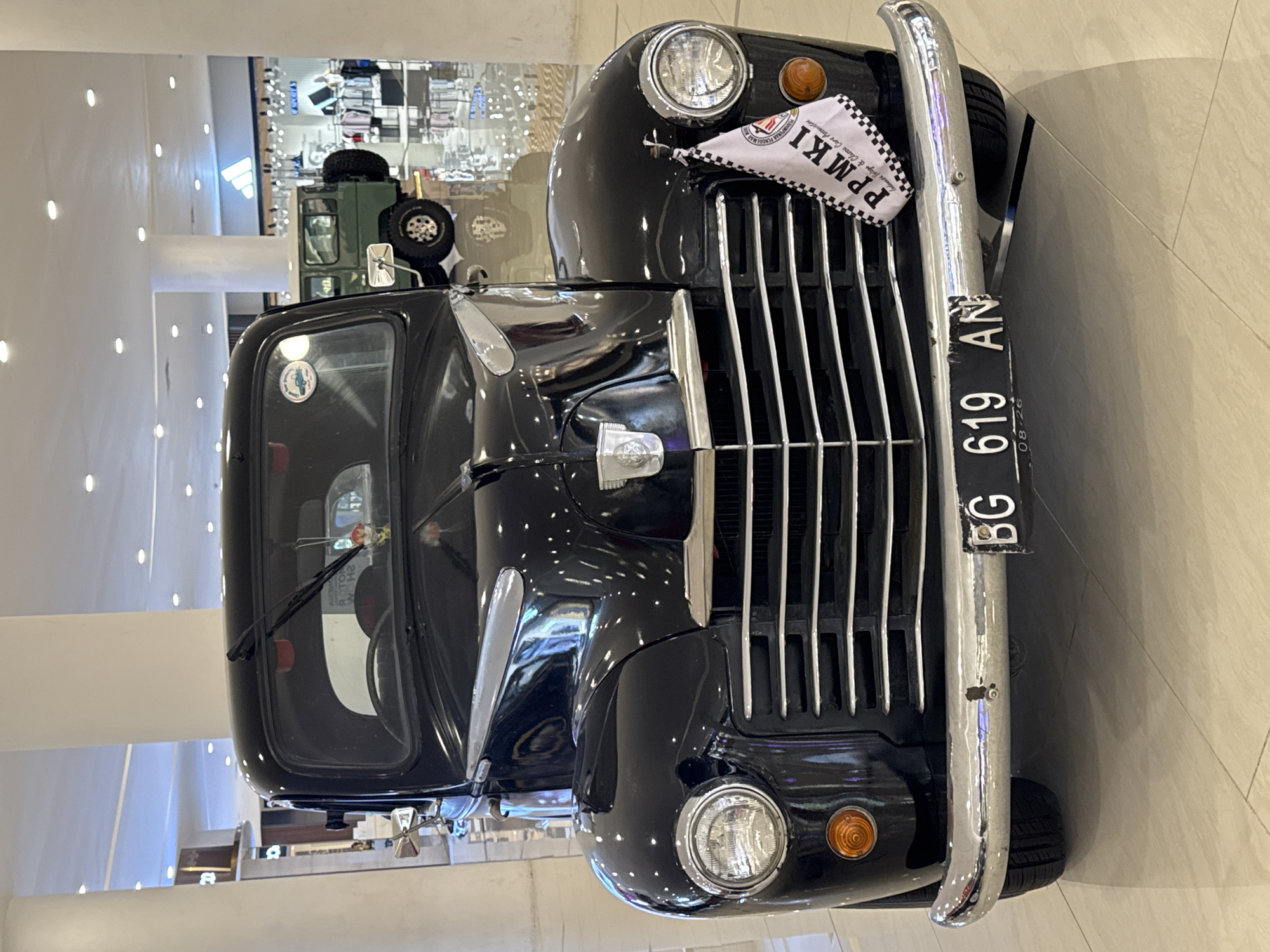 A black vintage car, possibly a pickup truck, is displayed indoors in what appears to be a shopping mall. The car is facing the viewer, showing its front grill, headlights, and bumper. A license plate reading "BG 619 AN" is visible. A flag with "PPMKI" and a checkered pattern is attached near the headlight. In the background, an army green vehicle, possibly a jeep, is partially visible behind the black car. The overall environment suggests a display or exhibit within a commercial setting, likely in the city of Kuta, Indonesia, based on the car's license plate and likely location. The foreground is a light-colored tiled floor. The mall setting is indicated by various retail displays and lighting fixtures. No people are visible in the image.