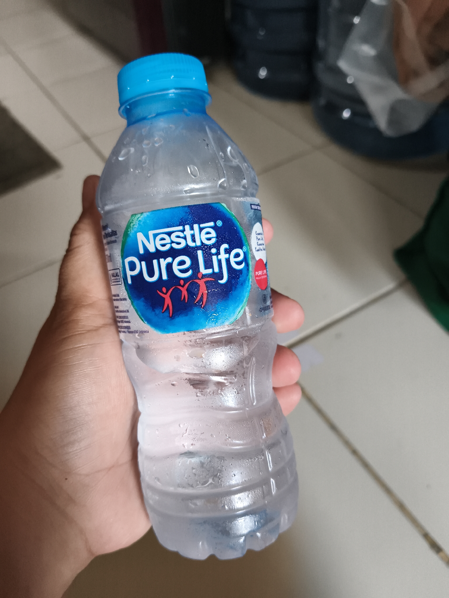 A hand holds a condensation-covered bottle of Nestle Pure Life water. The bottle has a blue cap and a blue and white label featuring the Nestle logo and stylized red figures. The background consists of a tiled floor and blurred large water jugs. The location is identified as Tangerang, Indonesia.