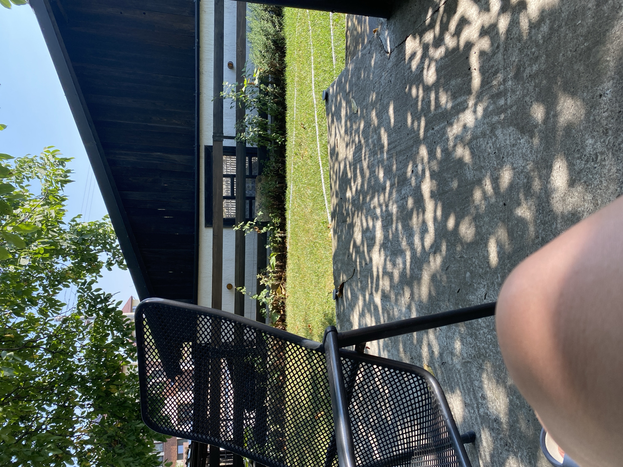 The image depicts an outdoor scene likely located in Târgu Neamț, Romania. In the foreground, a person's leg and knee are visible, with a grey metallic outdoor chair positioned to the left. The chair's mesh seat is partially in shadow. Beyond the chair and the concrete ground, which is dappled with sunlight and tree shadows, is a well-maintained lawn. Behind the lawn, a building with dark wooden siding on the upper part and white horizontally striped walls below is visible. A black horizontal railing with a darker wooden fence or structure behind it is in front of the building. Green plants and shrubs are growing in front of the building. Lush green tree foliage partially obscures the sky and upper portion of the building. The overall impression is of a serene, sunny day in a backyard or patio area.