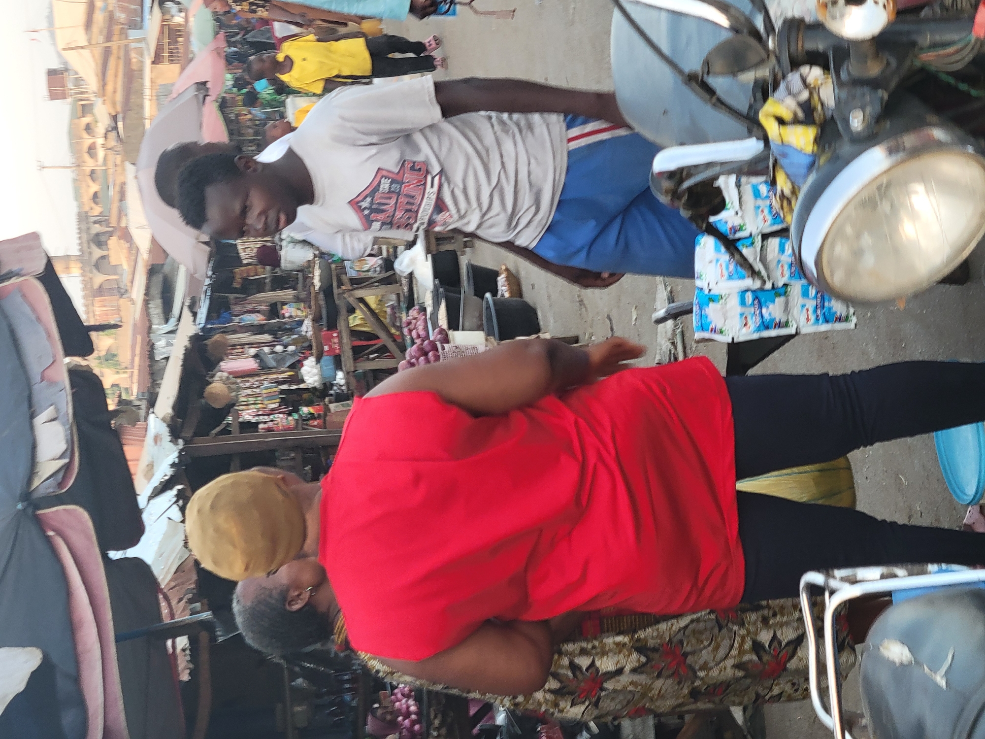 The image captures a busy street scene in Madalla, Nigeria, with several people visible. A young man wearing a white T-shirt and blue pants stands near a motorcycle loaded with small packets. An older woman with a yellow head covering and wearing a red shirt sits nearby. A younger man in a yellow shirt is seen in the background. The market environment is cluttered with various goods and structures.