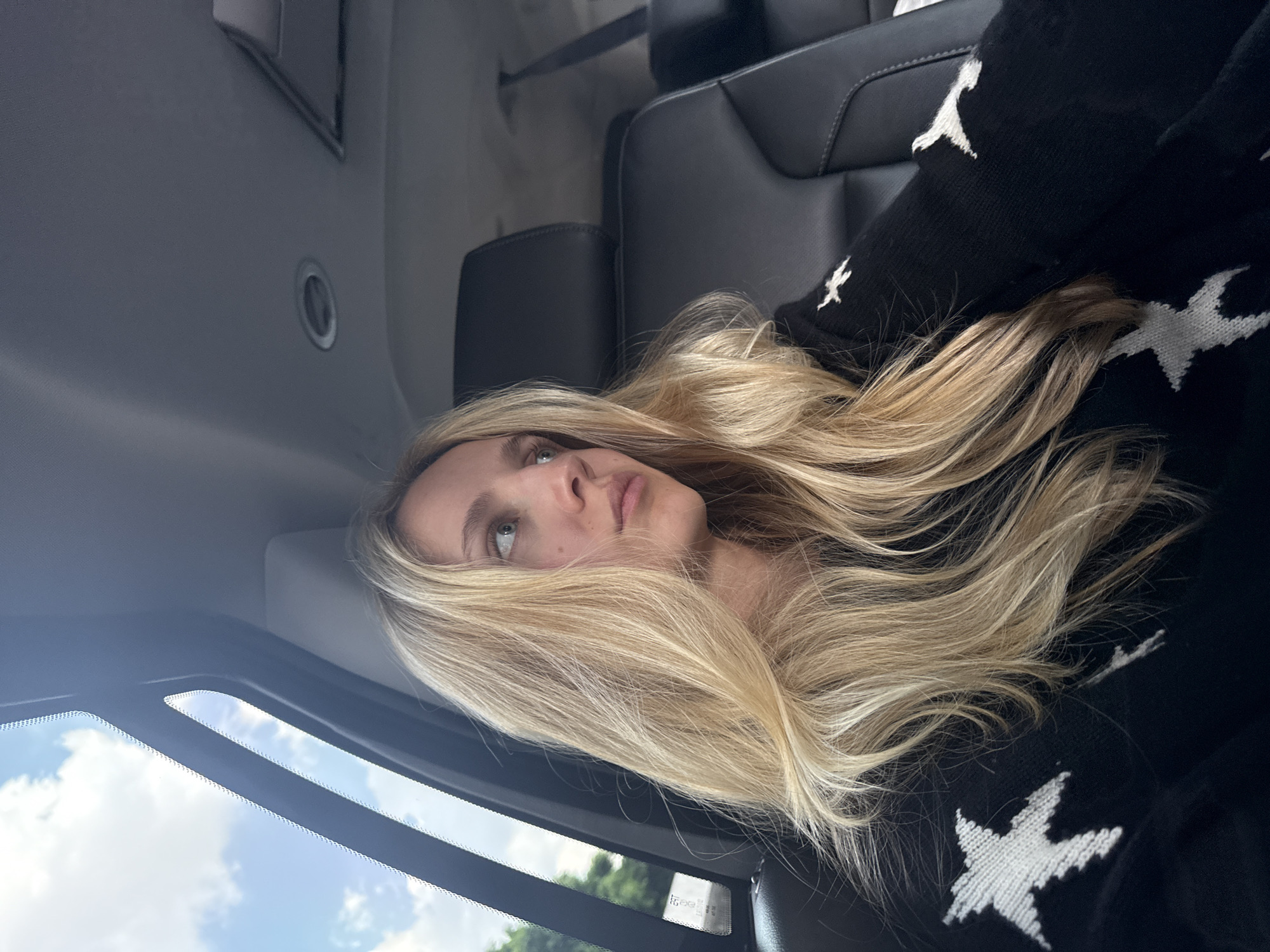 A young Caucasian female with long blonde hair is seated in the back of a car, looking out the window. She is wearing a black sweater with white stars. The interior of the car is visible, with black leather seats and a ceiling with lights. Outside the window, a cloudy sky and green trees are visible. The image is taken from a slightly high angle, looking down at the woman. It's not possible to determine the specific location within Mexico City from the image.