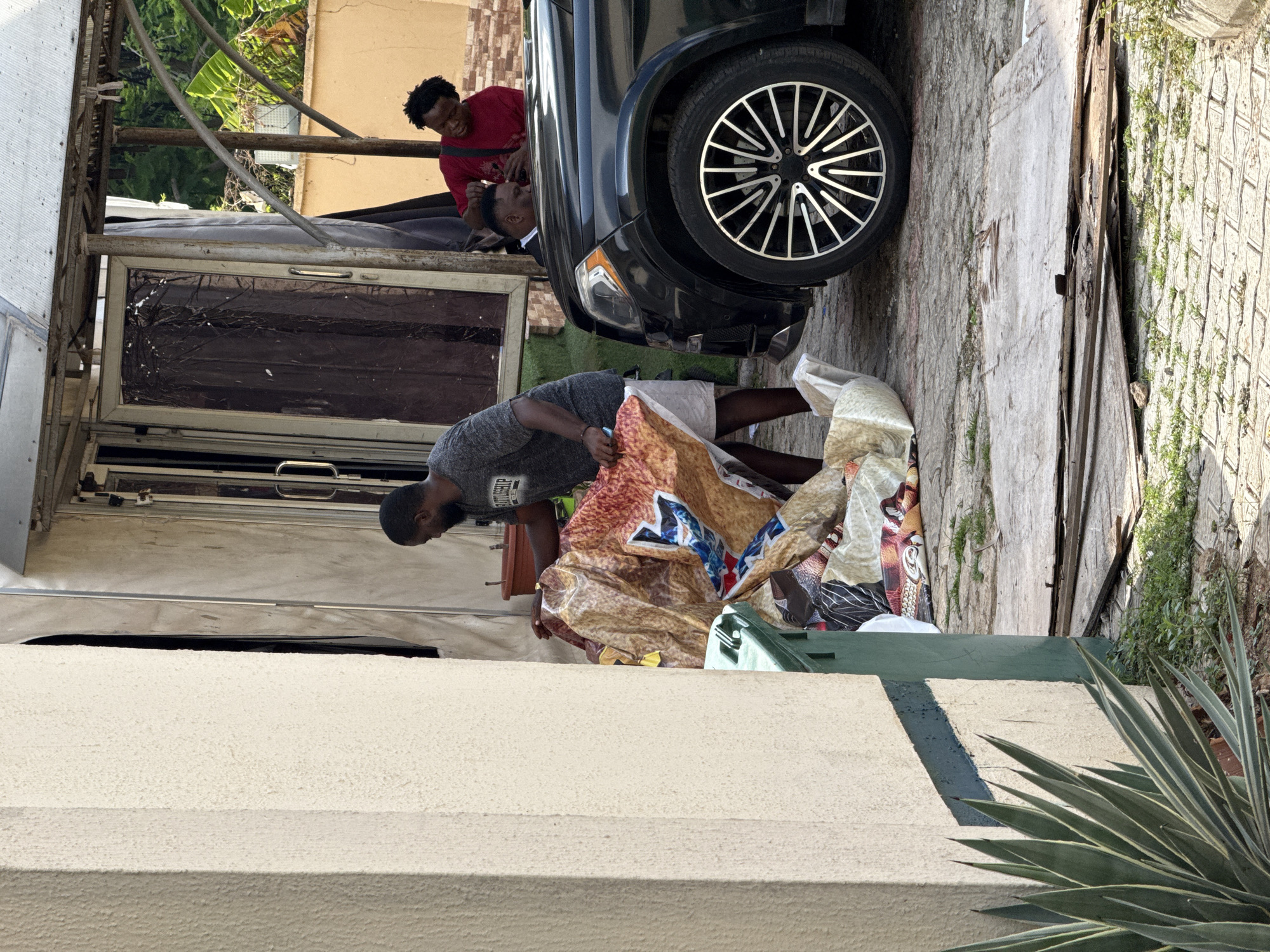A man in a gray t-shirt and white shorts is unloading items wrapped in patterned cloth from the back of a black car, specifically the tire area of the car. Two men are standing near the car. One man is wearing a red t-shirt. The scene takes place outside a building with a large window, a wall, and some plants. The location is inferred to be in Abuja, Nigeria.