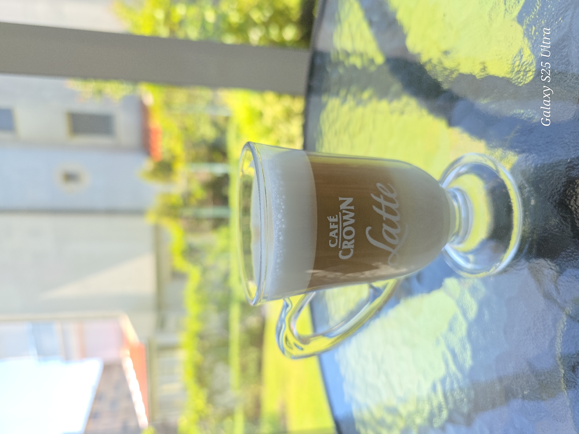 A latte in a glass with the words "CAFÉ CROWN Latte" printed on it is on a textured glass table. In the background is a building with a window and a lush green outdoor area with flowers. The image was taken with a Galaxy S25 Ultra and it is speculated that the location is Sveti Vlas, Bulgaria.