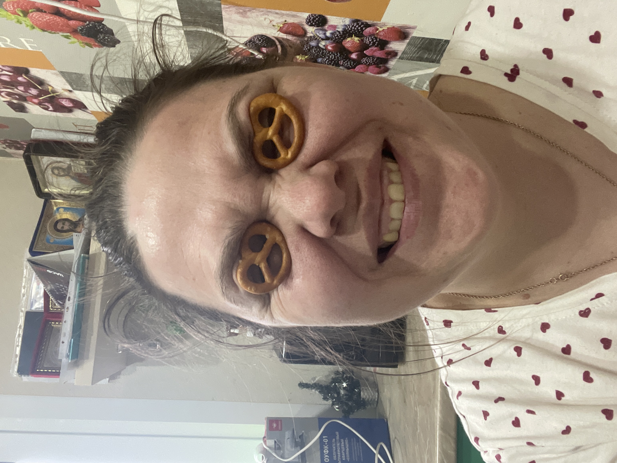 The image, rotated 90 degrees counter-clockwise, depicts a woman playfully posing indoors in a home setting in Noviy, Russia. She is smiling broadly with a cheerful expression, and has small, hard pretzels placed over each of her closed eyes, creating a humorous effect. Her dark hair is somewhat messy, and she is wearing a white t-shirt adorned with small red heart patterns. A thin gold chain is visible around her neck.
The background reveals a domestic environment. The wall behind her is covered with wallpaper featuring a pattern of various fruits, including strawberries, blackberries, and cherries. To her left, two framed religious icons are visible on a shelf or surface, suggesting a Christian household. In the lower part of the image, below her head, a small, artificial Christmas tree with tiny decorations can be seen. Next to it is a blue and white box with Cyrillic text.
Visible text on the blue box reads "ОУФК-01" and "Облучатель кварцевый КВАРЦЕВЫЙ «СОЛНЫШКО»" (Quartz Irradiator "Sun"), indicating a Russian-made device, consistent with the specified location. Part of the word "RE" is also visible on the wallpaper. The lighting suggests an indoor setting, likely during the daytime or with artificial light. The overall mood of the image is lighthearted and joyful.