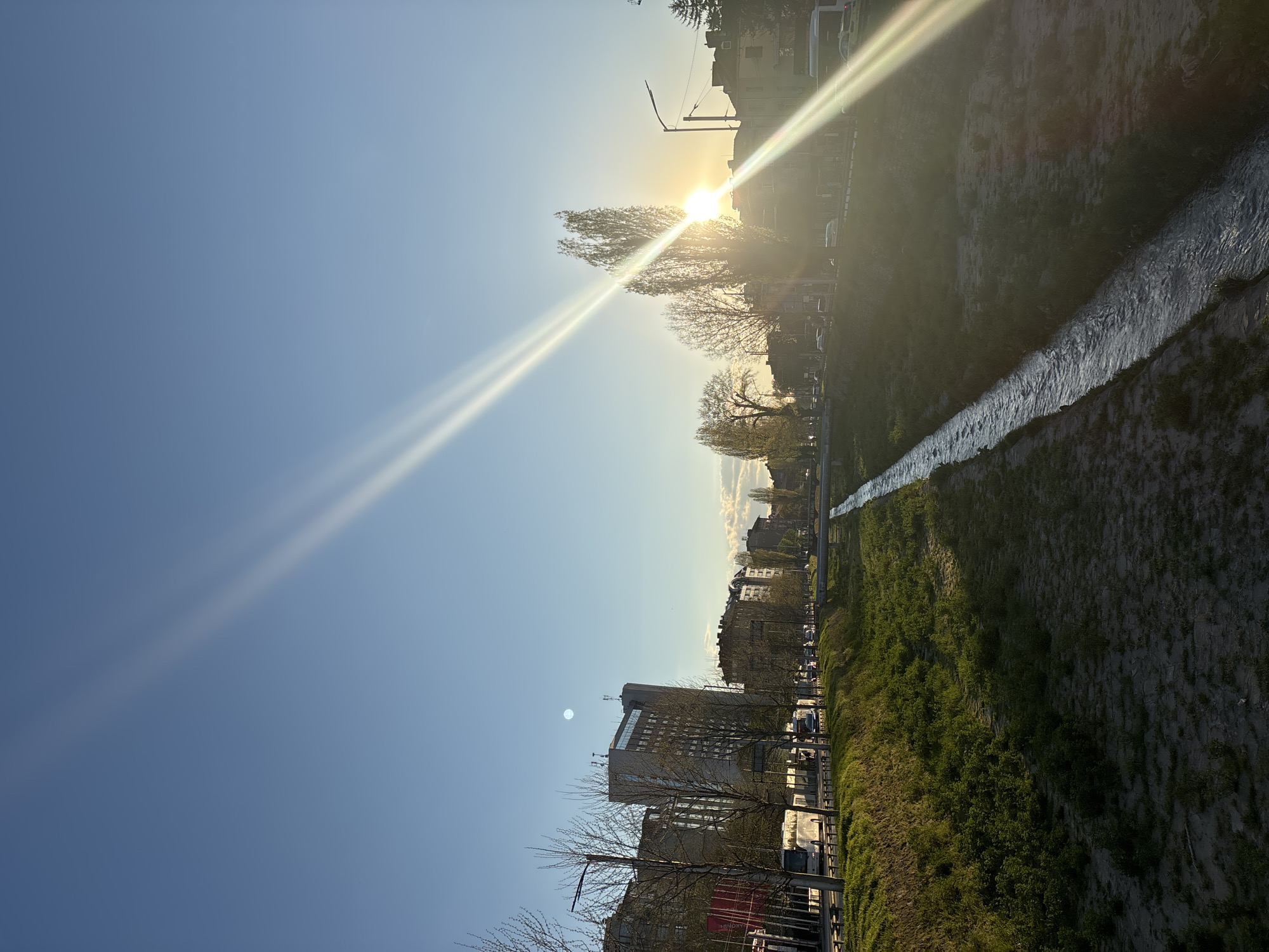 The image shows a cityscape with a river flowing through it. The location is Sofia, Bulgaria, identifiable by the characteristic buildings and the canalization of the Perlovska River. The sun is setting or rising, creating strong glare and light streaks across the sky and landscape. A row of buildings lines the left side of the image, while the river runs along the right, confined by concrete banks. Green grass grows along the riverbank, and trees are scattered throughout the scene. Cars can be seen parked along the street next to the river. The sky is clear and blue. The orientation of the photo is rotated from vertical to horizontal.
