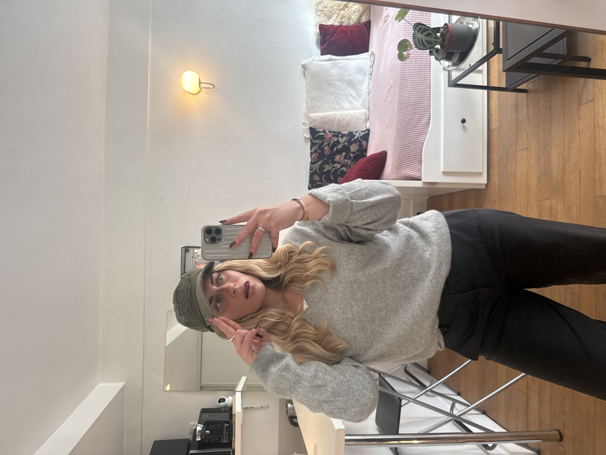 A young woman stands in front of a mirror taking a selfie with her phone. She's wearing a grey sweater, black pants, and a green hat. Her blonde hair is styled in waves. In the background, a bed with multiple pillows, a small bedside table with a plant, and a light fixture on the wall are visible. The scene is indoors, likely in an apartment in Paris, France. A chair is also visible. A coffee maker and security camera is seen in the mirror on the lower left side. The floors are hardwood and the walls are white.