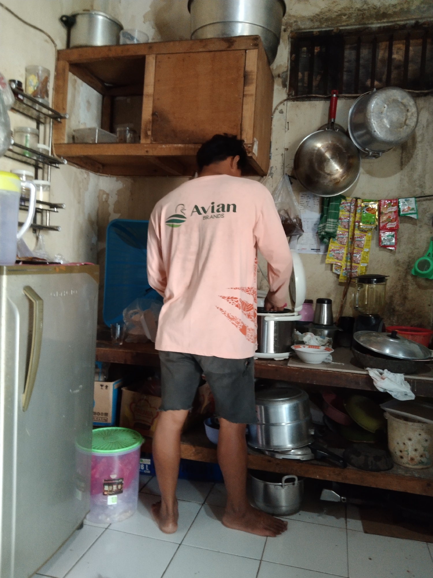 The image depicts an indoor kitchen scene in Parung, Indonesia. A person, viewed from the back, is wearing a light pink long-sleeved shirt with an "Avian Brands" logo and dark shorts. They are standing in front of a counter laden with various kitchen items. To the left, a silver refrigerator stands tall. The kitchen walls are a pale, somewhat grimy color. Shelves above the counter hold pots, pans, and various containers. To the right, more shelves are stocked with packaged goods, a blender, and more cookware. A large, green-lidded plastic container is visible near the refrigerator. The overall impression is one of a functional, lived-in kitchen with ample storage and a utilitarian aesthetic.