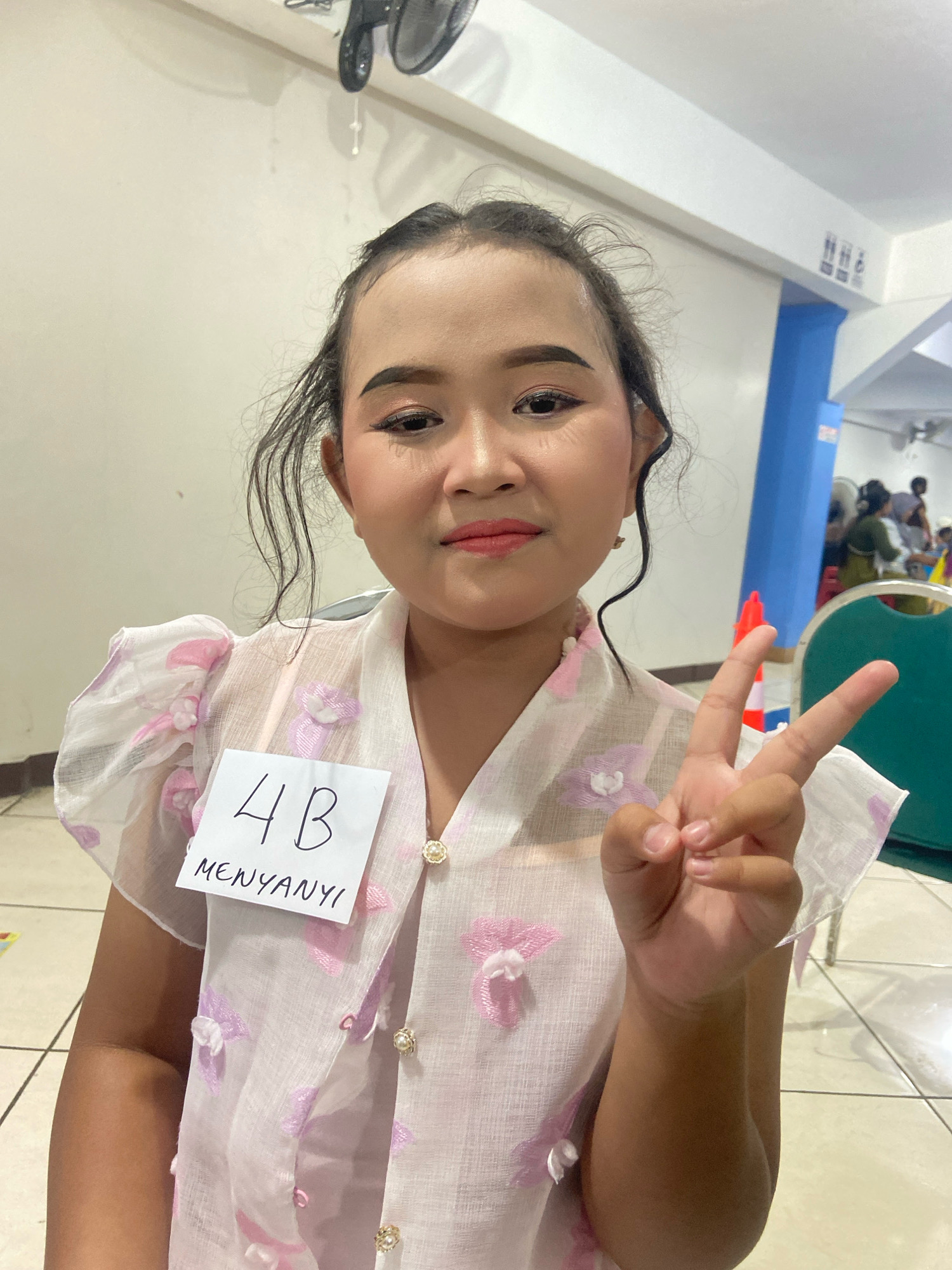 The image shows a young girl in the city of Kecamatan Pati, Indonesia, posing for a photo. She is wearing a light pink top with floral embellishments and has a nametag attached to her shirt that reads "4B MENYANYI". The girl is holding up two fingers in a peace sign. Her makeup is noticeable, with defined eyebrows, eyeliner, and lipstick. The background appears to be an indoor space with plain walls, a ceiling fan, and a blue column. There are other people and some orange cones visible in the background, suggesting an event or competition is taking place.