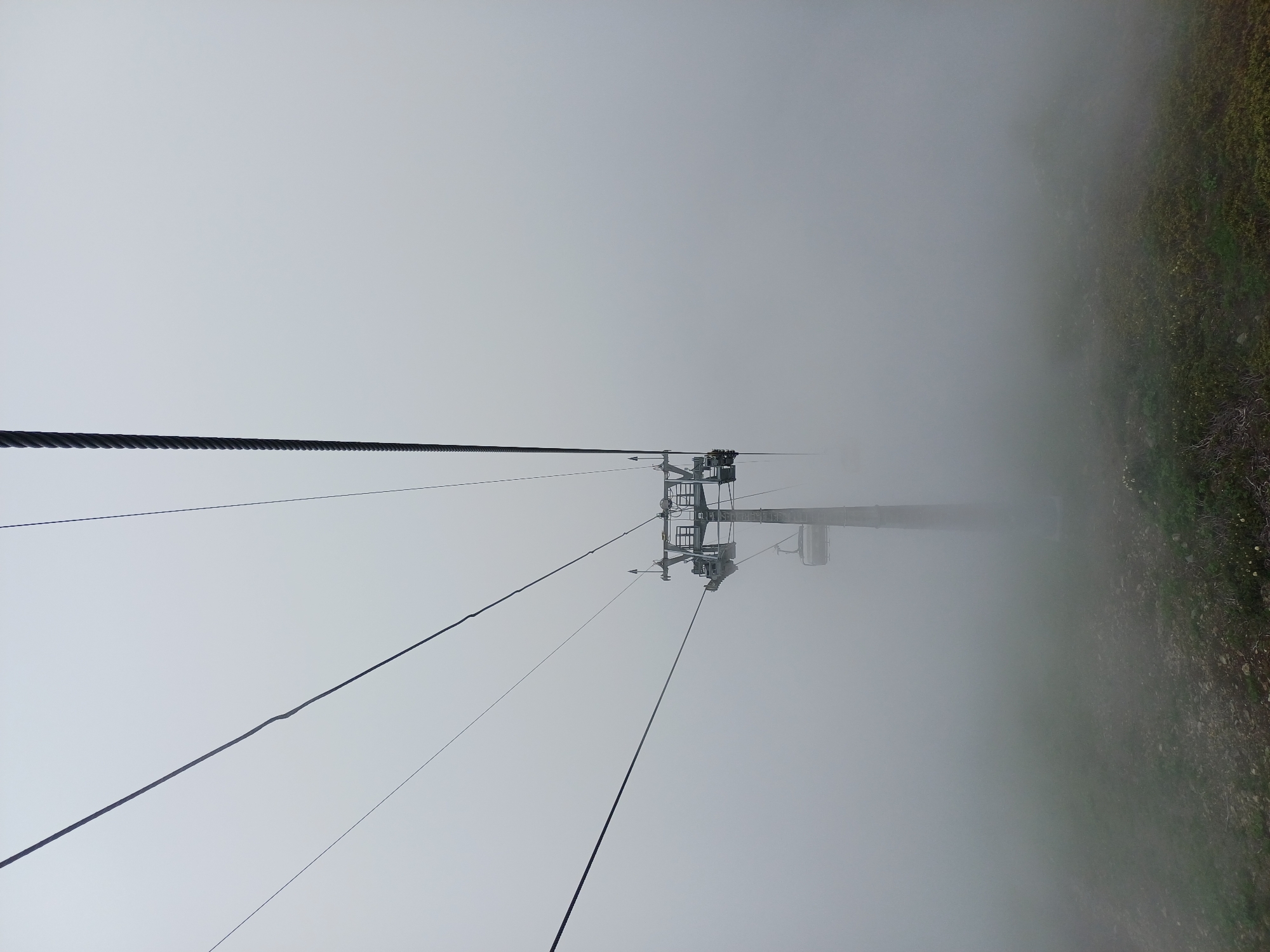 An overcast, foggy day obscures a ski lift tower and cables in Estosadok, Russia. The ski lift structure, made of metal, stands tall and is partially obscured by thick fog. Several cables are visible stretching across the frame, disappearing into the mist. The ground in the foreground is covered in sparse vegetation and rocks. The overall atmosphere is muted and damp due to the fog, typical of a cloudy mountain environment.