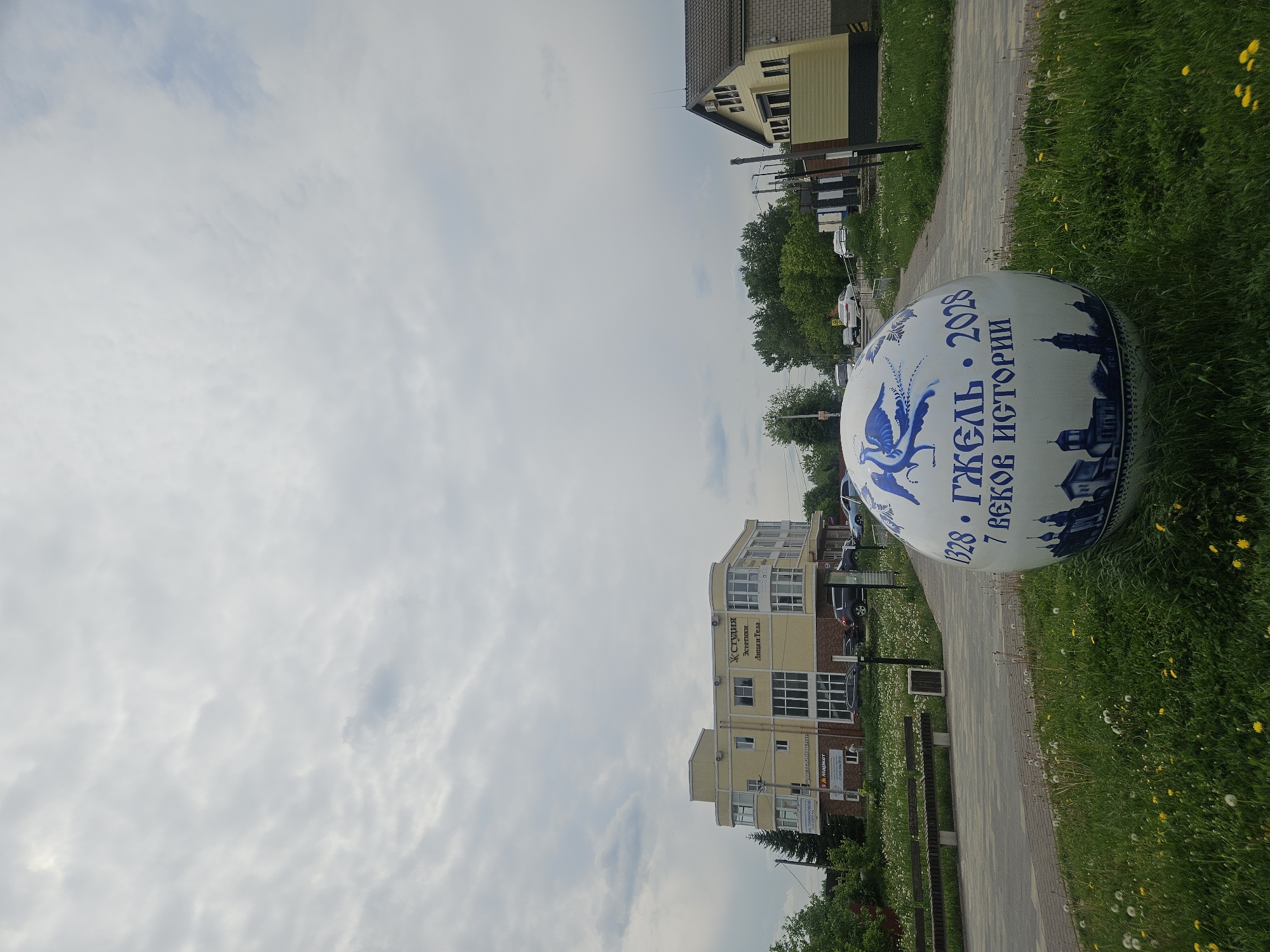 The photo shows an exterior scene in Речицы, Russia, featuring a large, spherical decorative object placed beside a paved walkway. The object is white with blue accents, depicting a stylized bird and the inscription "1328 • ГЖЕЛЬ • 2028, 7 ВЕКОВ ИСТОРИИ" in Russian.
The walkway is edged by grass, which contains dandelions. In the background, there are buildings, including a light-yellow building with the inscription "Студия Эстетики Лица и Тела" (Studio of Aesthetics for Face and Body). Also, there is a two-story house with light brown siding.
The sky is overcast with light clouds.