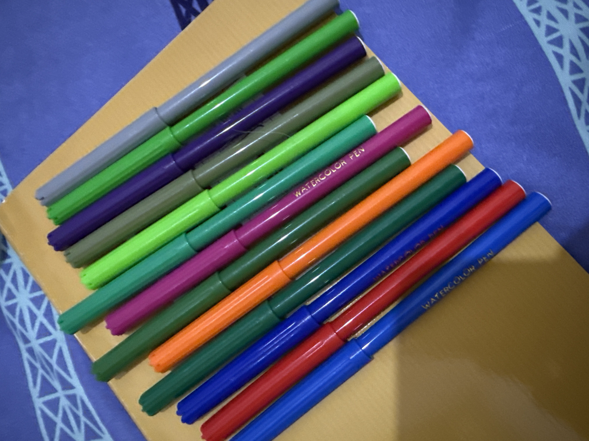 A close-up, angled shot shows a collection of multi-colored marker pens arranged diagonally on a golden-yellow surface, which appears to be a notebook or portfolio. The markers are predominantly in shades of green, purple, orange, red, and blue, with a few gray ones interspersed. The marker caps are visible, and some of the pens have "WATERCOLOR PEN" printed on them in white text. The golden surface rests on a blue fabric with a repeating geometric white pattern. The lighting suggests an indoor setting, possibly a desk or table. There are no people or discernible actions taking place, it is a still life composition of art supplies. The location of Pematang Siantar, Indonesia, is not identifiable from the visual information present in the image.