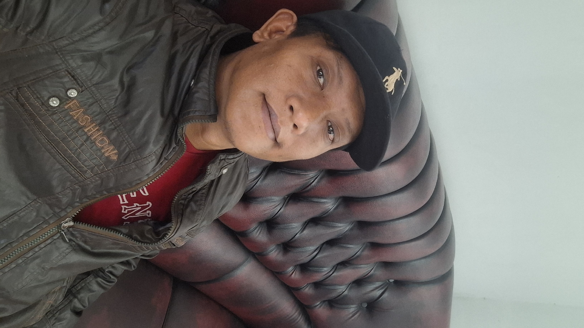 A man wearing a black baseball cap with a gold logo and a brown jacket over a red shirt is seated on a tufted brown leather couch. The man is looking directly at the camera with a slight smile. The background is a plain light-colored wall. The scene appears to be indoors, possibly in a home or a lounge area. The image quality is fair, with some graininess and slight blurriness. The location is Ciharashas, Indonesia.