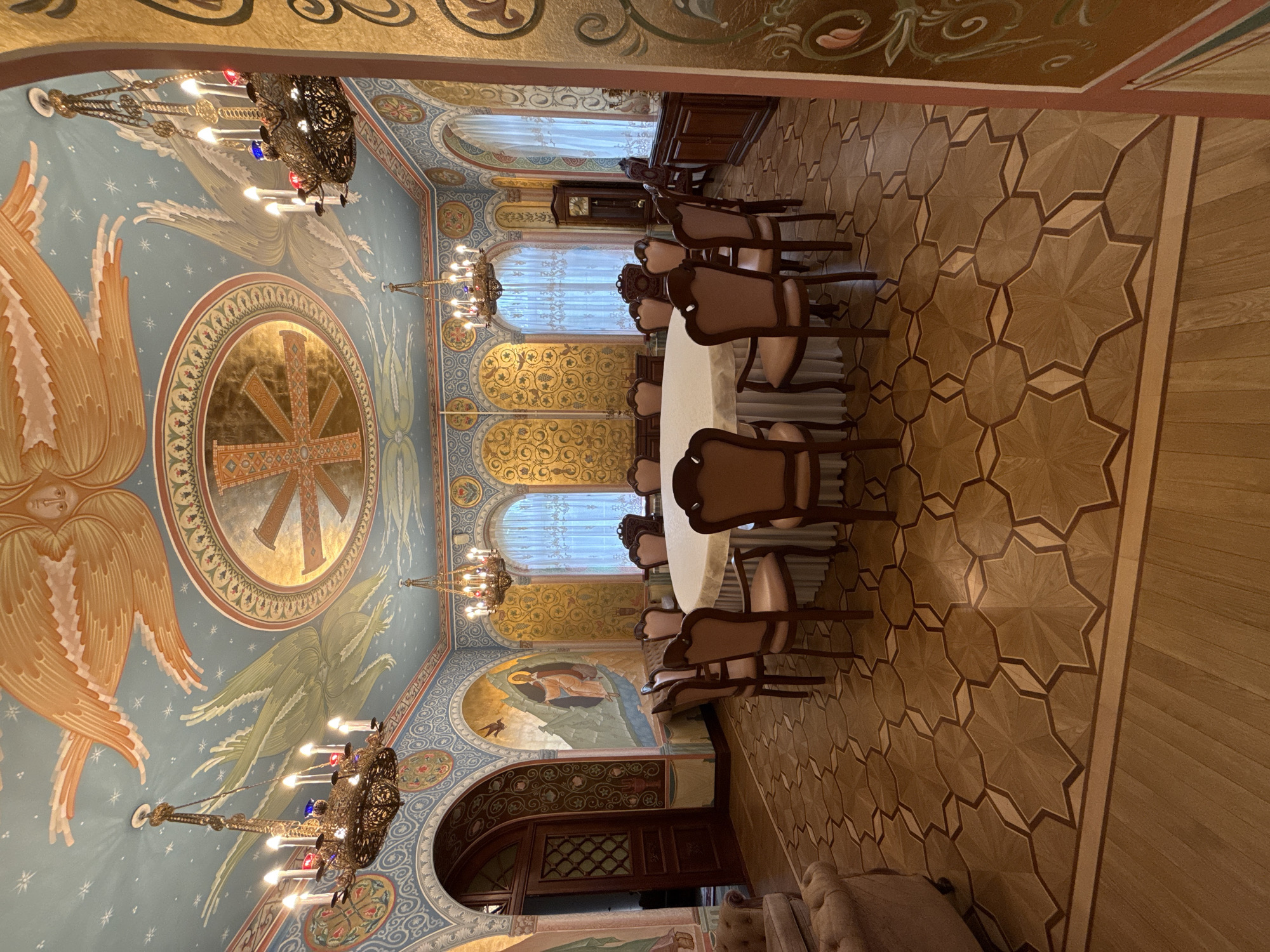 The image shows the interior of the Church of the Savior on Spilled Blood in Saint Petersburg, Russia. The room features a large round table covered with a white tablecloth, surrounded by wooden chairs. Above, the ceiling is elaborately decorated with religious iconography, including angels and a prominent cross within a circle. Chandeliers hang from the ceiling. The walls are adorned with murals and decorative patterns. The floor is covered with patterned wooden flooring. There are arched windows with curtains and what appears to be a cabinet and grandfather clock in the background. An arched doorway is visible at the end of the room.